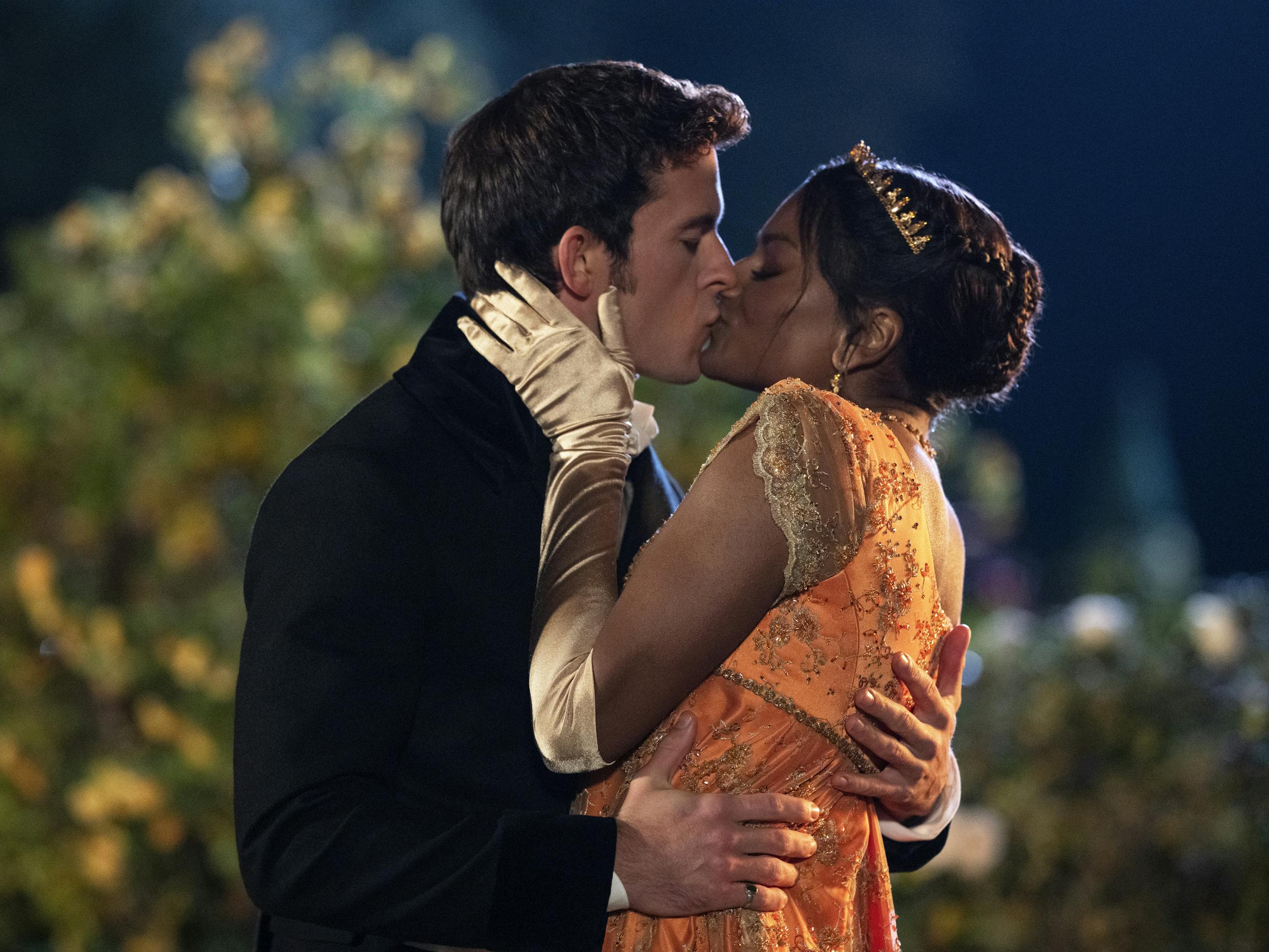 Anthony Bridgerton (Jonathan Bailey) and Kate Sharma (Simone Ashley) kiss in an outdoor scene. She wears an orange dress and tiara, and he wears a dark suit.