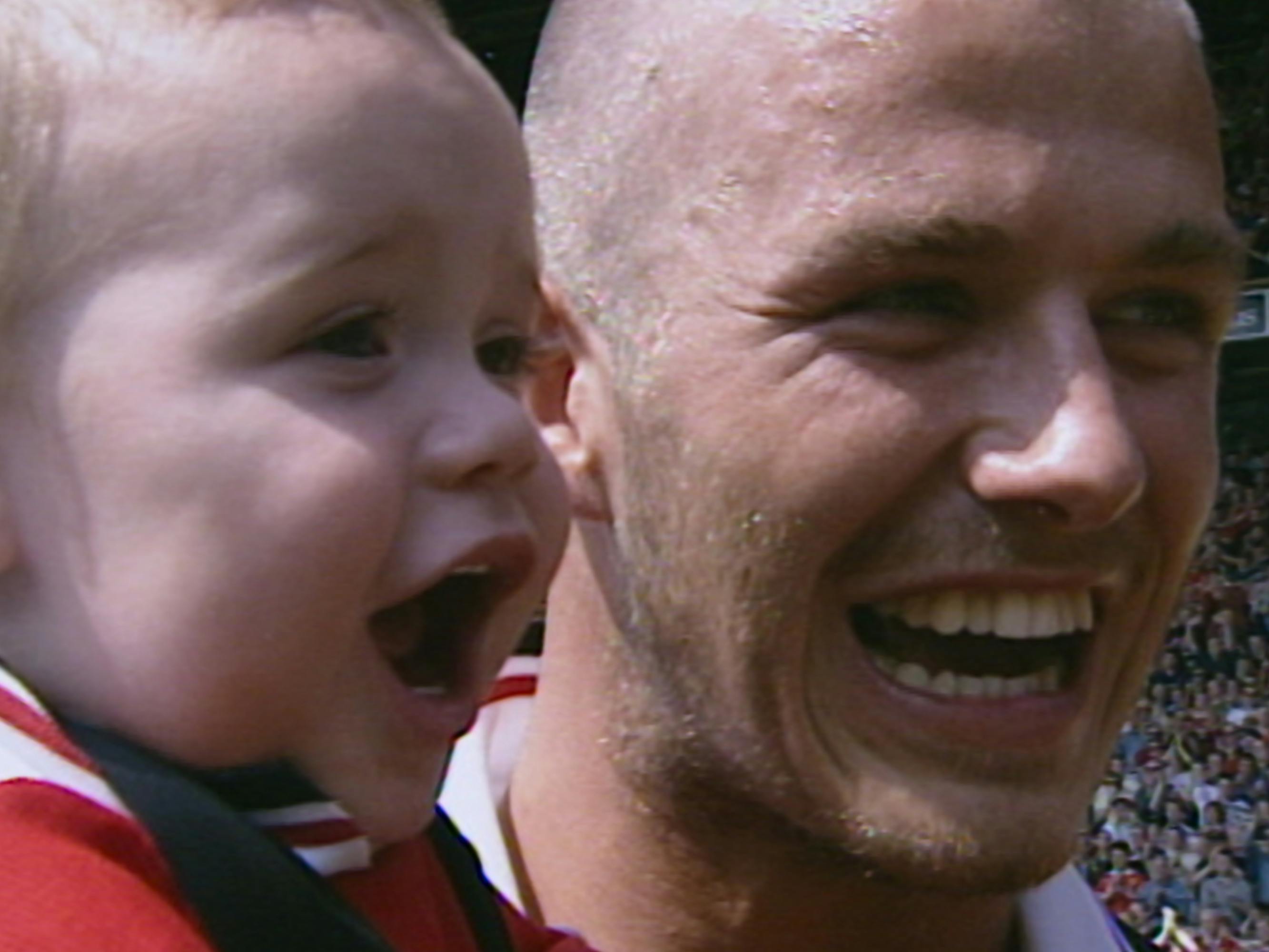 David Beckham holds Brooklyn Beckham in a crowded soccer pitch. Both Beckhams cheer with wide-mouthed smiles, and baby Brooklyn wears white earplugs and looks downright cherubic.