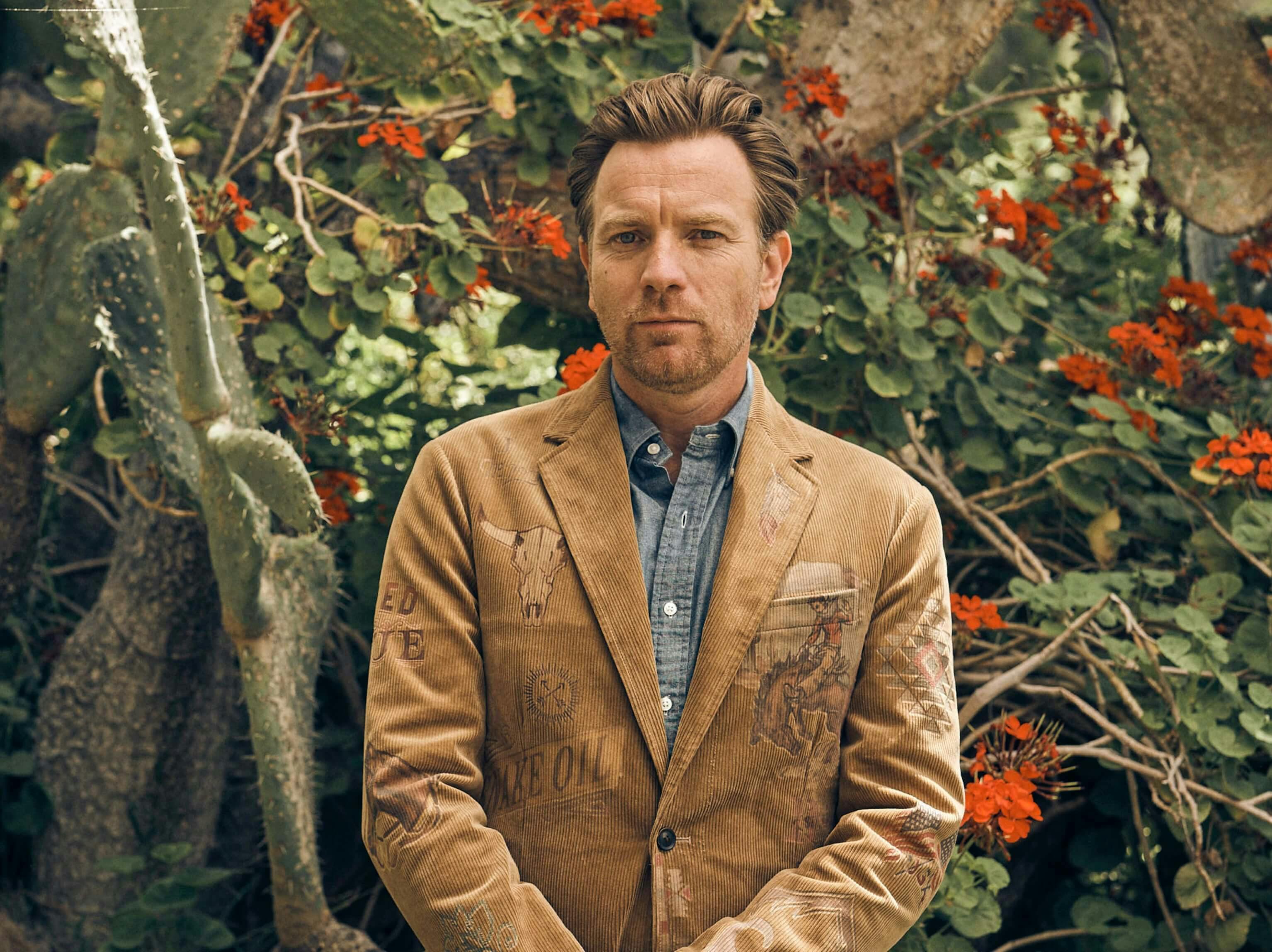 Ewan McGregor stands in front of a bush with red flowers and green leaves. He wears a tan coat and a grey shirt beneath. His hair is slicked back and he looks discerningly at the camera. 