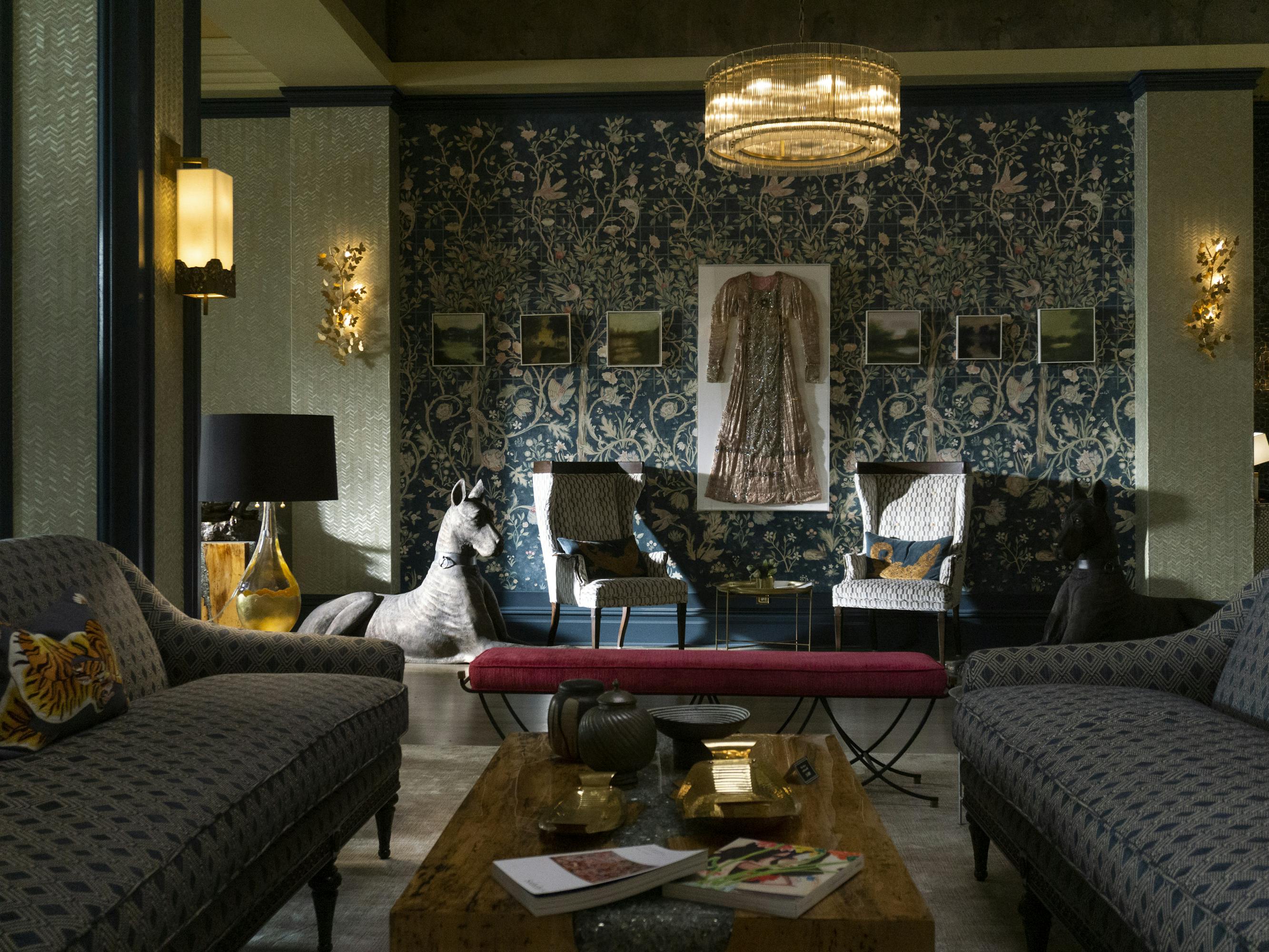 The 12 George Hotel. There are grey patterned couches, a red ottoman, a wooden coffee table, a grey creature in the top left corner, a black lamb, a dress hanging on the wall, art, and light fixtures.