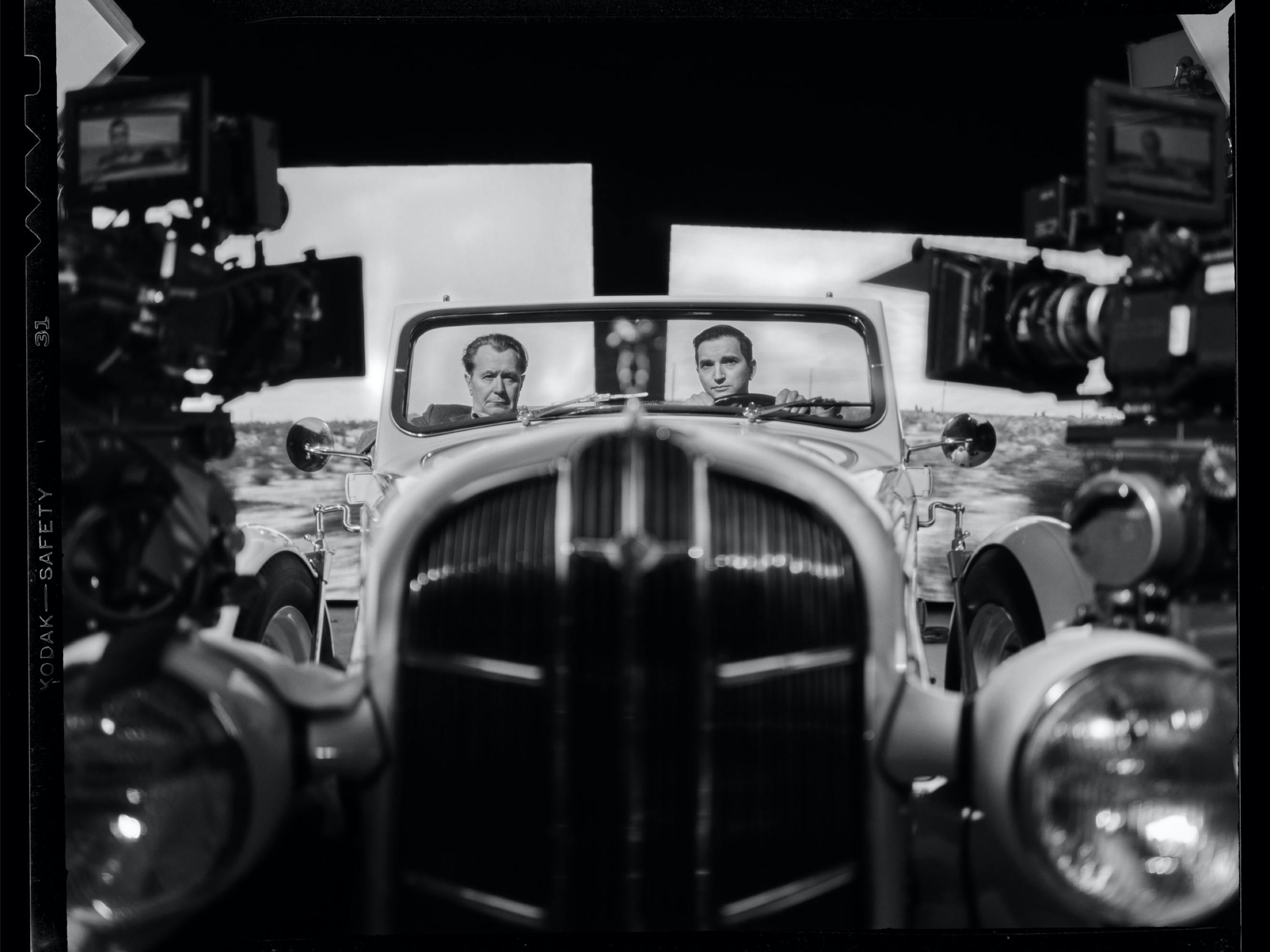 In this satisfyingly symmetrical black-and-white photo, Oldman and Persaud are visible from the head up behind the windshield of a sizable classic car. Behind them images of California scenery are projected on two screens. In front of them, we can see two cameras, each projecting out over one of the cars headlights