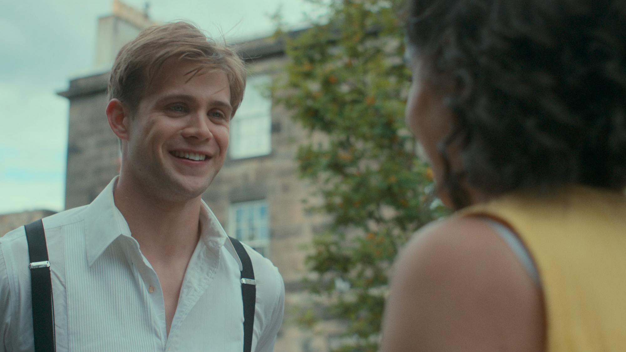 Dexter Mayhew (Leo Woodall) wears black suspenders over a white shirt and smiles at Emma Morley (Ambika Mod), whose back is turned from us. 