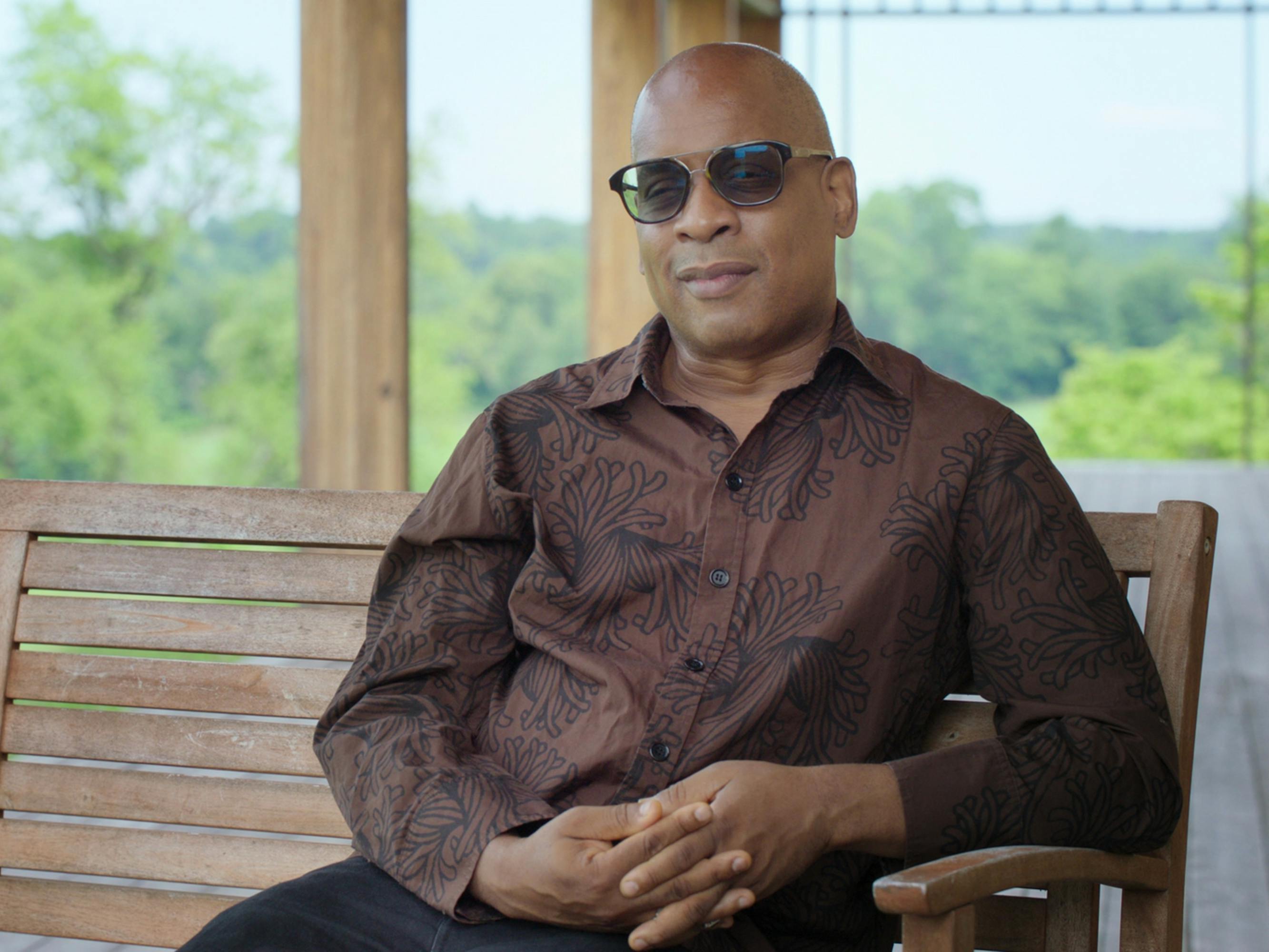 Glenn Ligon wears a brown shirt and shades and sits on a sunny porch, on a wooden bench.