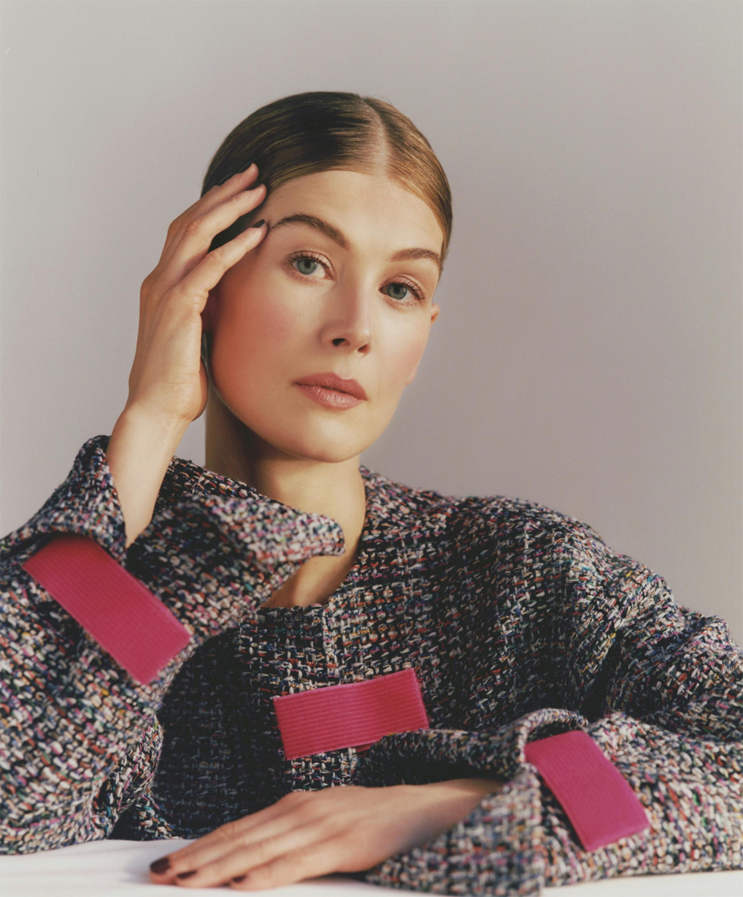 A portrait of Rosamund Pike wearing a tweed jacket with red accents on the cuffs and pocket. Her hair is pulled tightly back and parted down the center. She leans her head delicately on one hand.