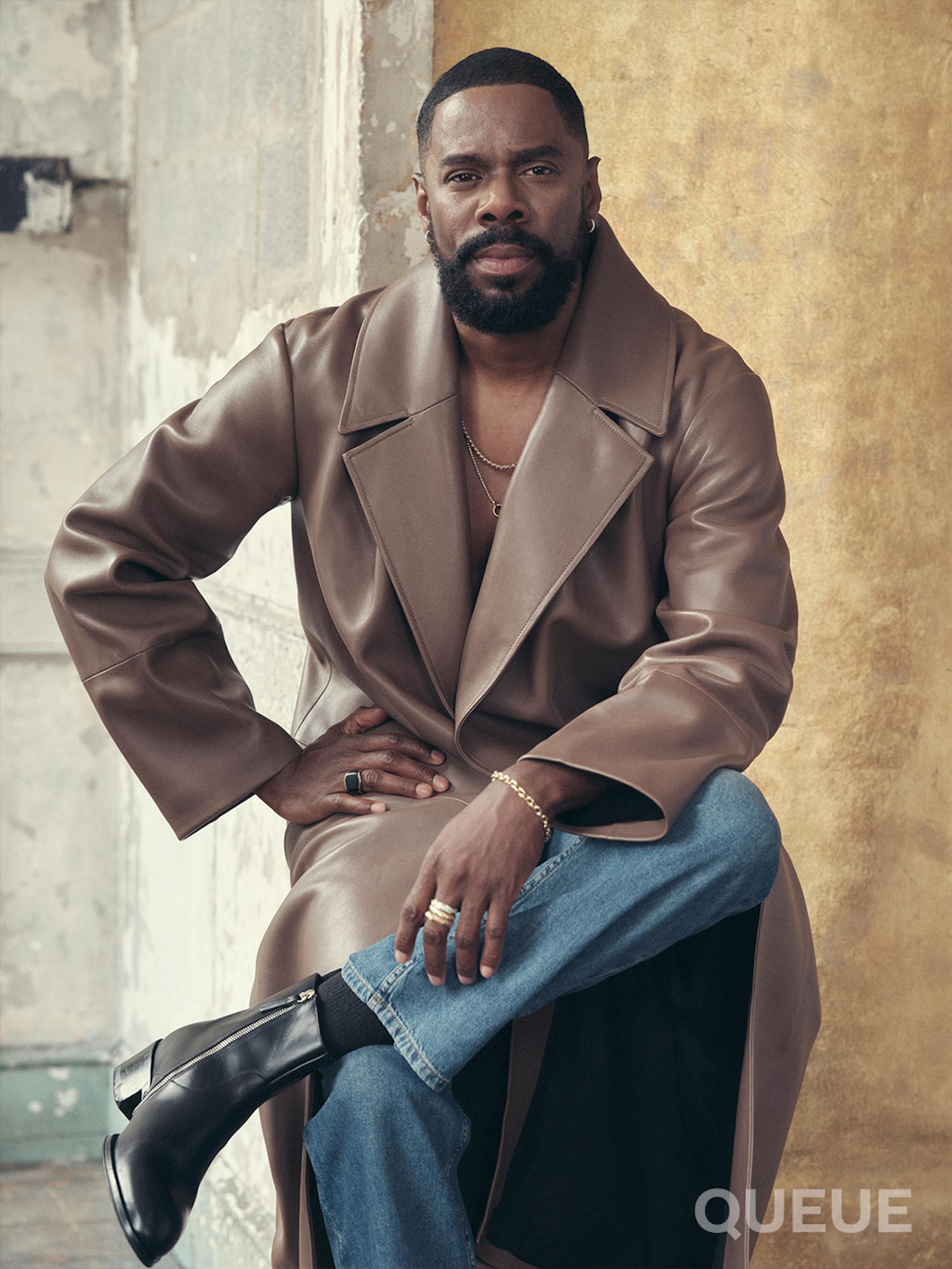 Domingo sits, wearing a brown leather trench coat, jeans, and black boots.