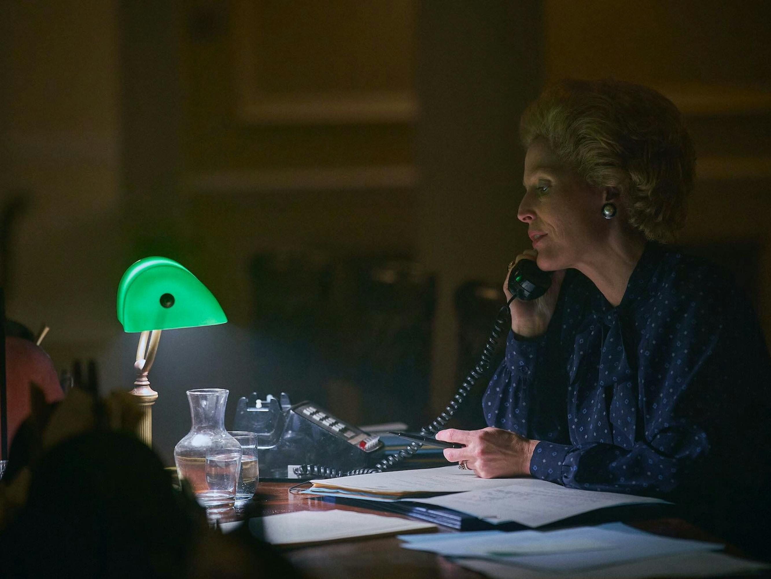 Margaret Thatcher (Gillian Anderson) talks to someone on the phone. She sits at a desk covered in papers, a bottle of water, and a green lamp. She wears a navy blouse.