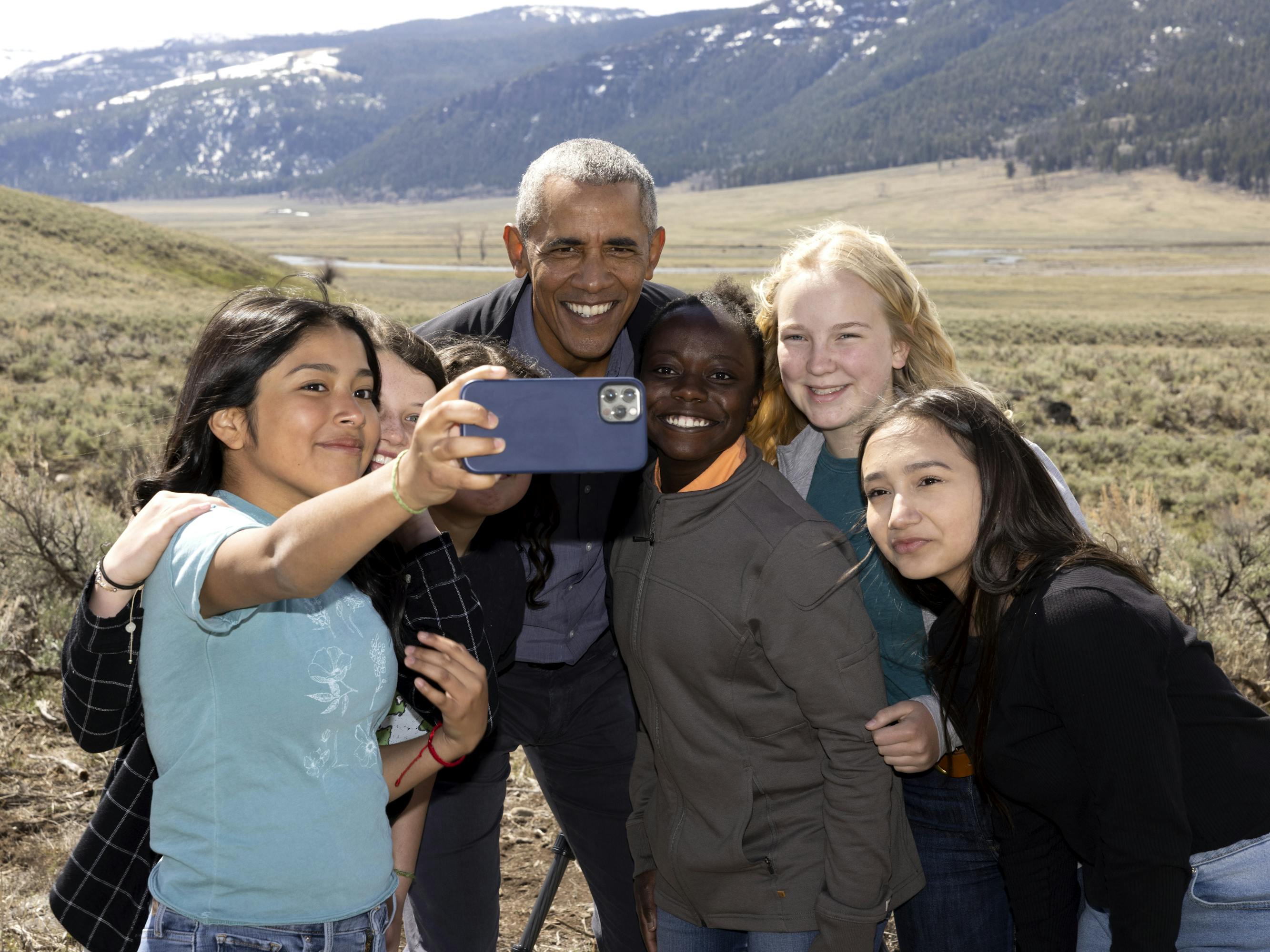Barack Obama takes a selfie with some people in a grassy expanse.