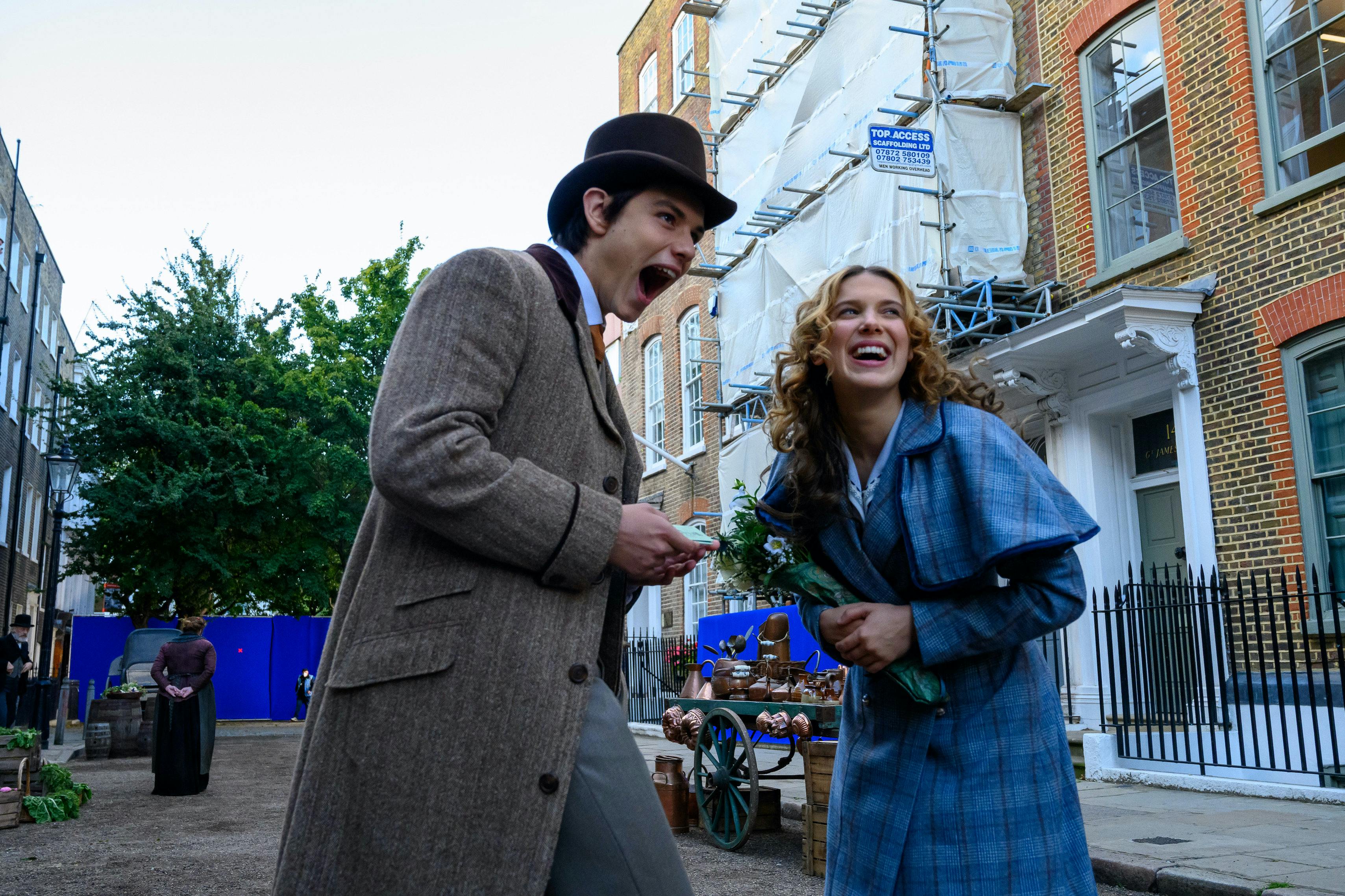 Tewkesbury (Louis Partridge) and Enola Holmes (Millie Bobby Brown) laugh in the street.