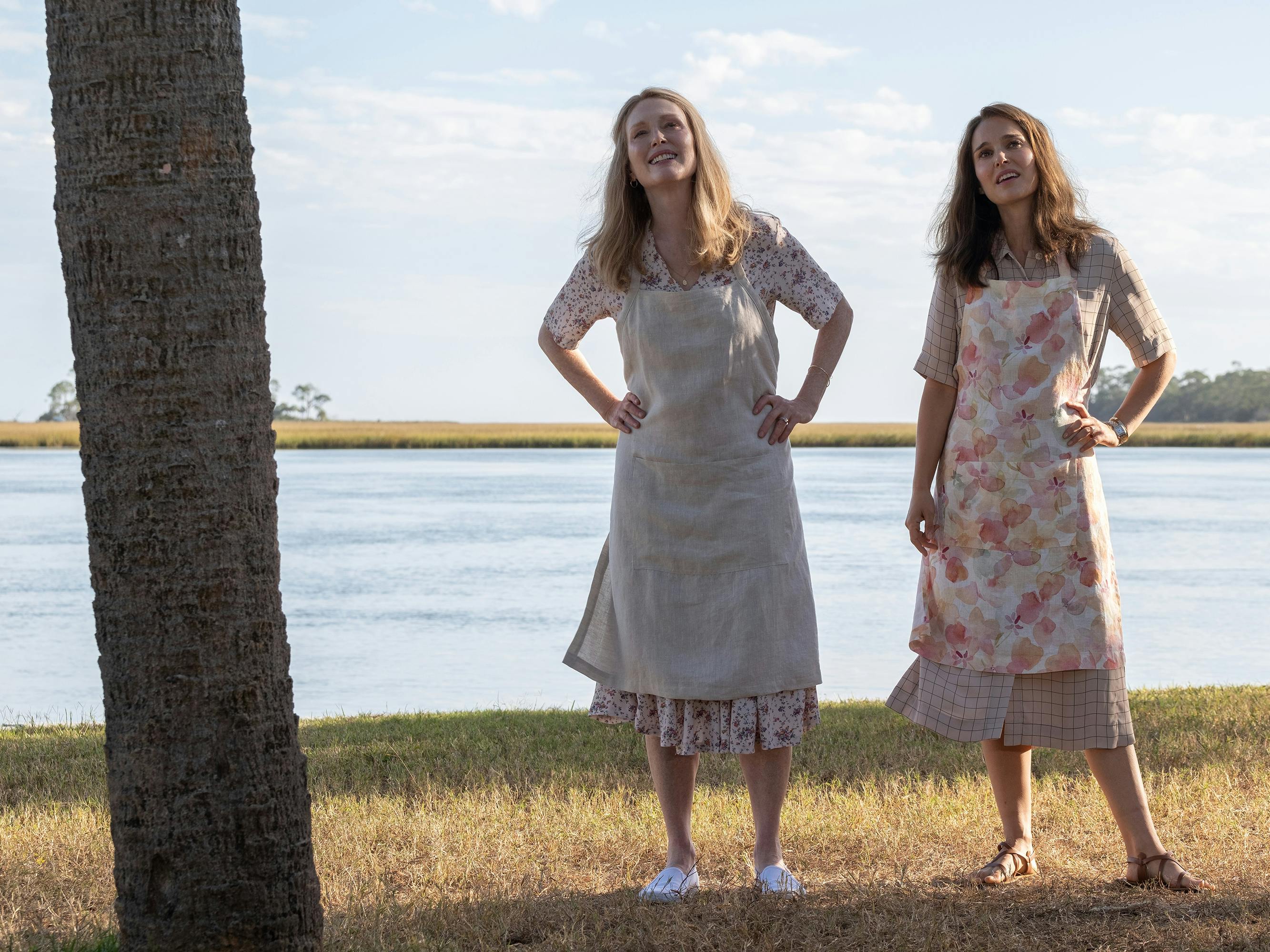 Gracie (Julianne Moore) and Elizabeth (Natalie Portman) stand together by a bay of water. Both wear aprons and midi-length dresses. They crane their necks looking at something in the sky.
