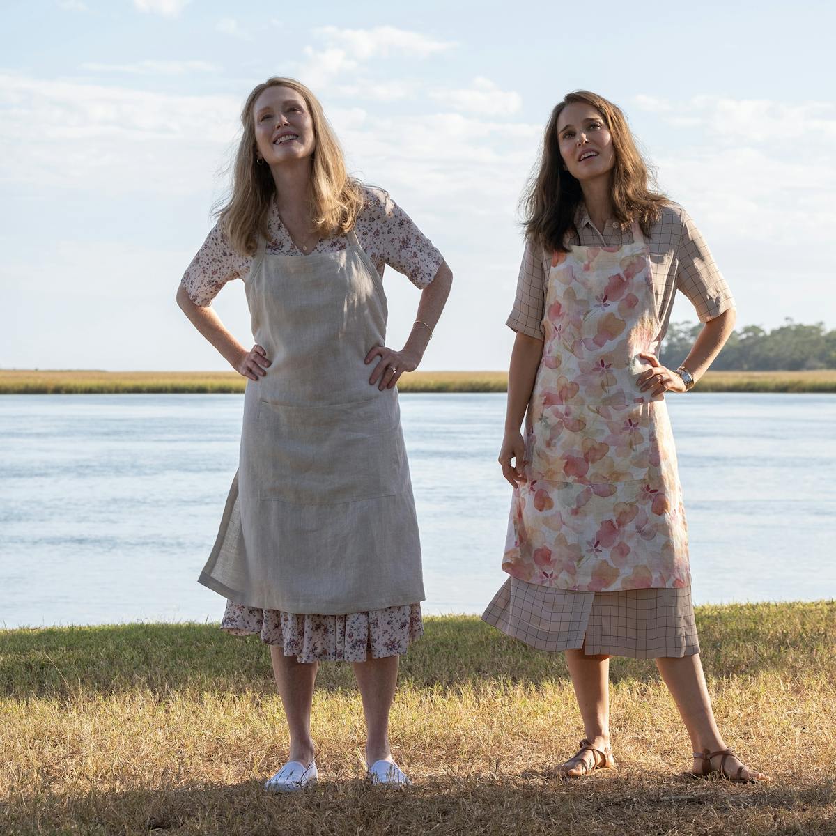 Gracie (Julianne Moore) and Elizabeth (Natalie Portman) stand together by a bay of water. Both wear aprons and midi-length dresses. They crane their necks looking at something in the sky.