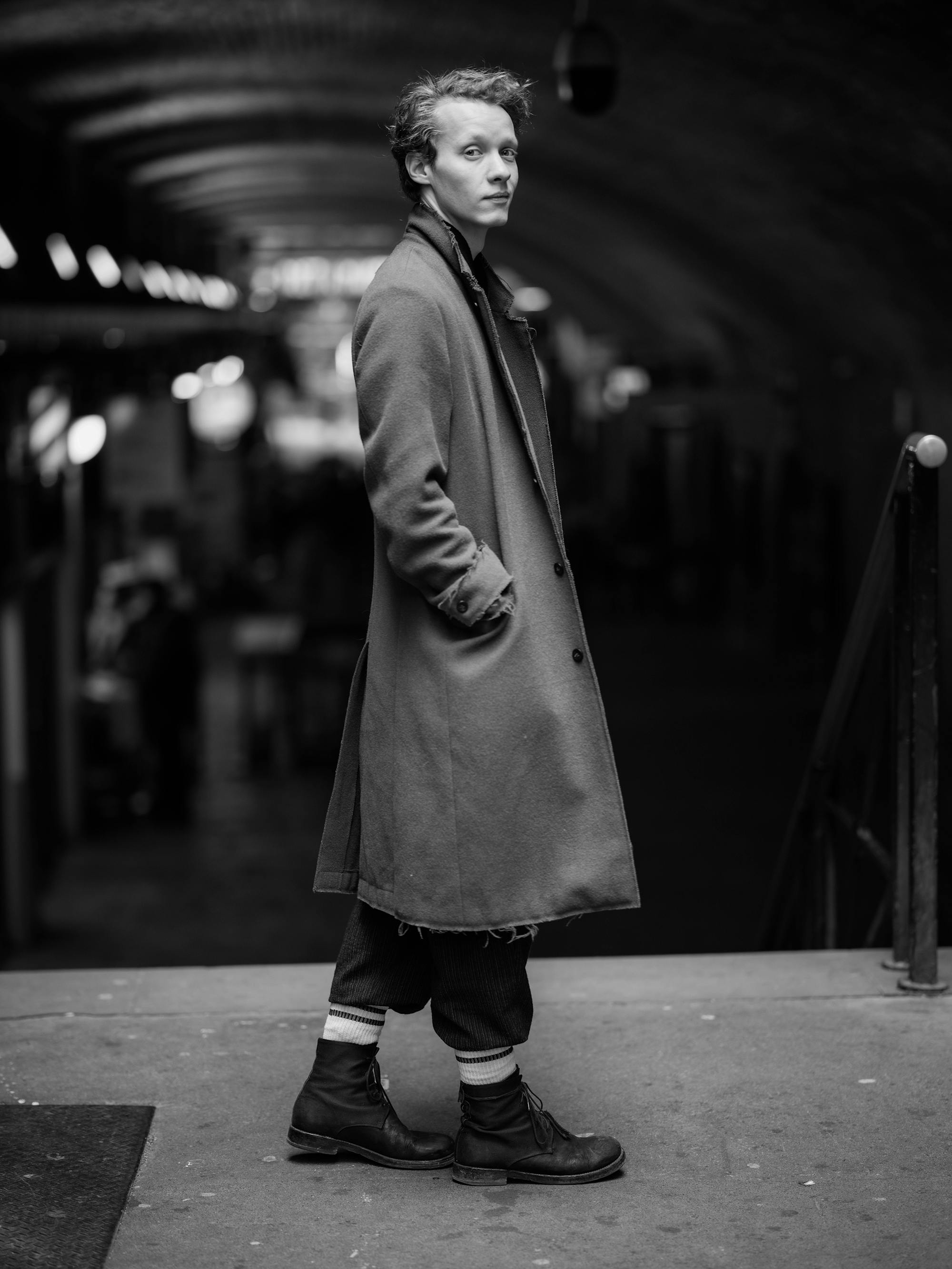 Felix Kammerer looks way cleaner than he does in the film. He wears a long coat, dark pants, and dark boots. He stands in what looks like a train station.
