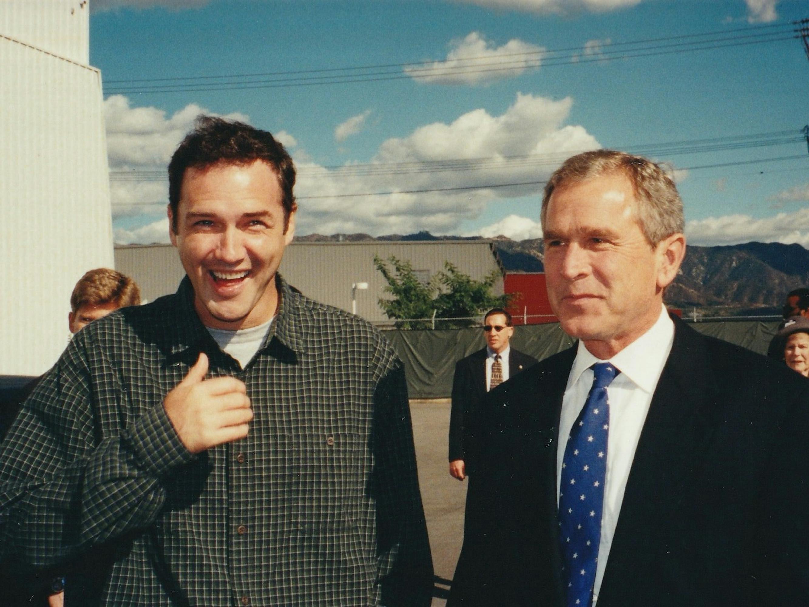 Norm Macdonald and George Bush stand together outside against a blue sky dotted with puffy clouds.