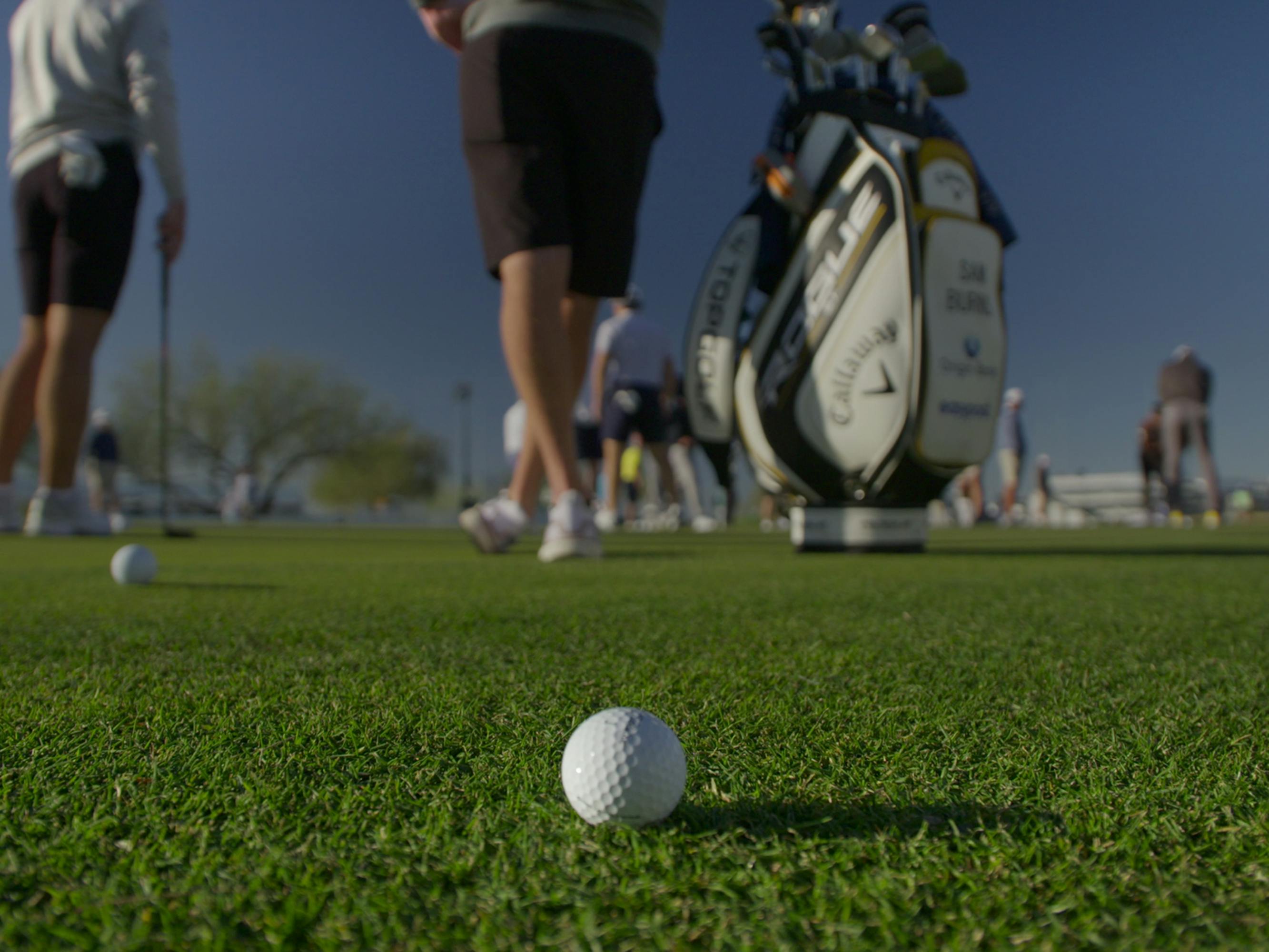 A golfball sits on the green. Behind the ball are some golfer's legs and a golf bag.