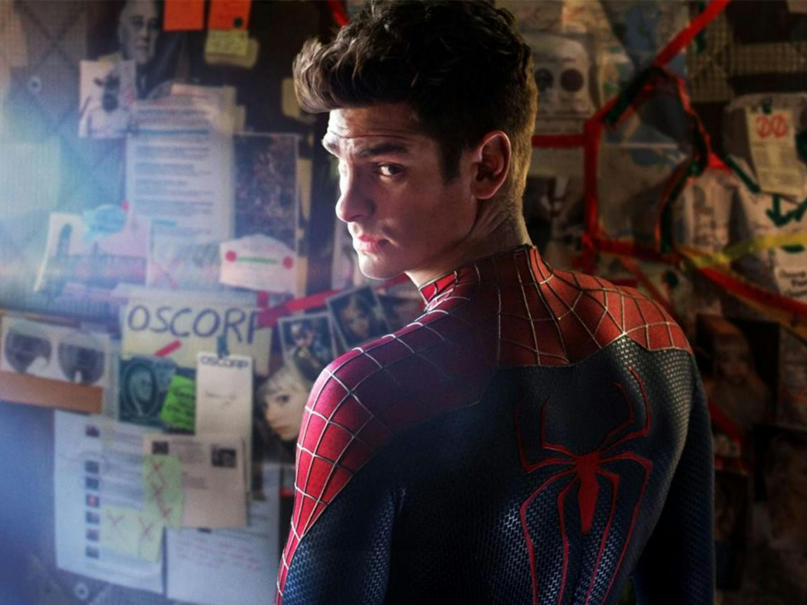 Andrew Garfield wears his spider-man outfit and stands in front of a wall covered with notices.