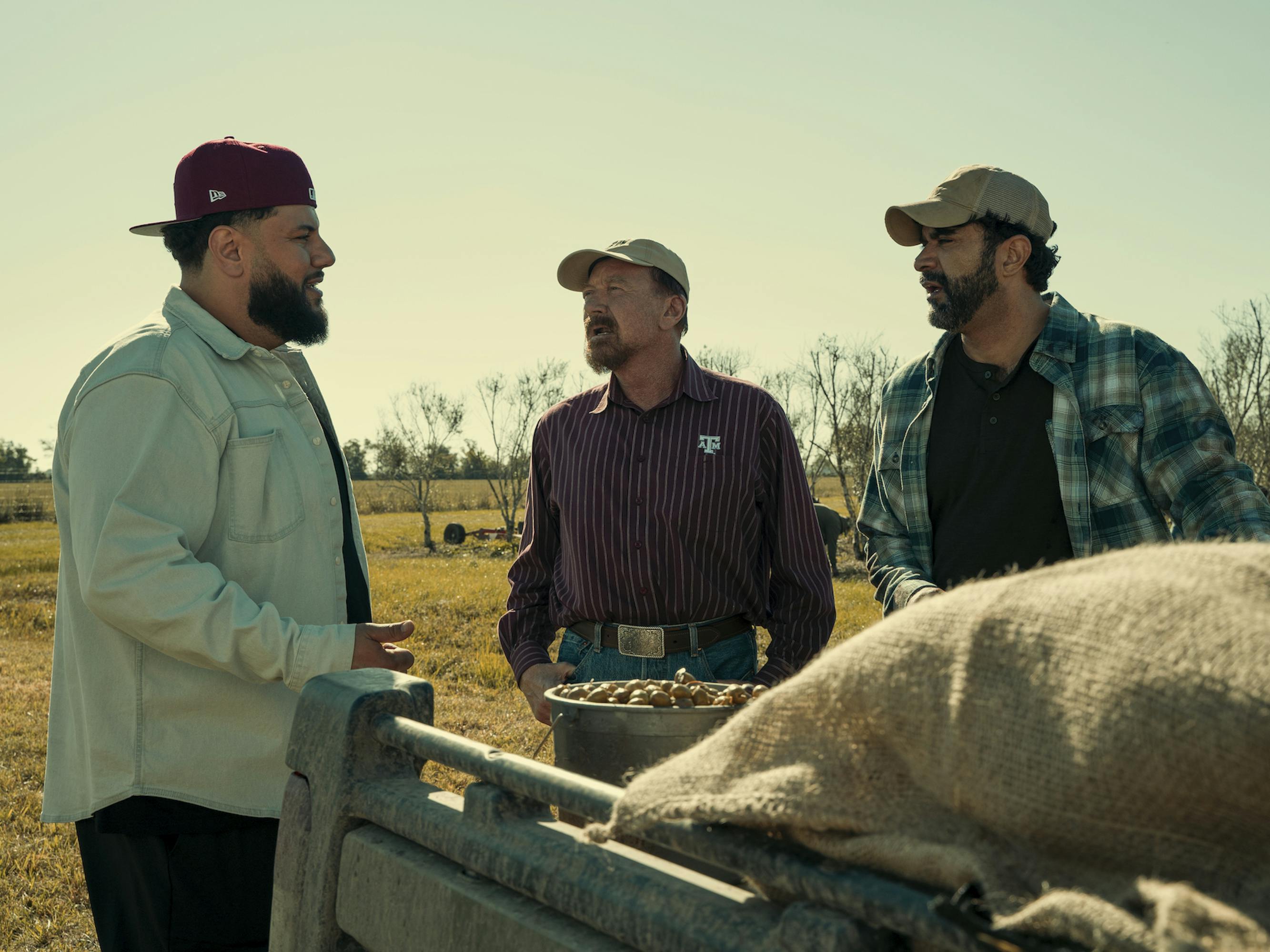 Mo (Mo Amer), Buddy (Walt Roberts), and Manny (Carlos Lerman) wear baseball hats and stand over a bin of a olives.