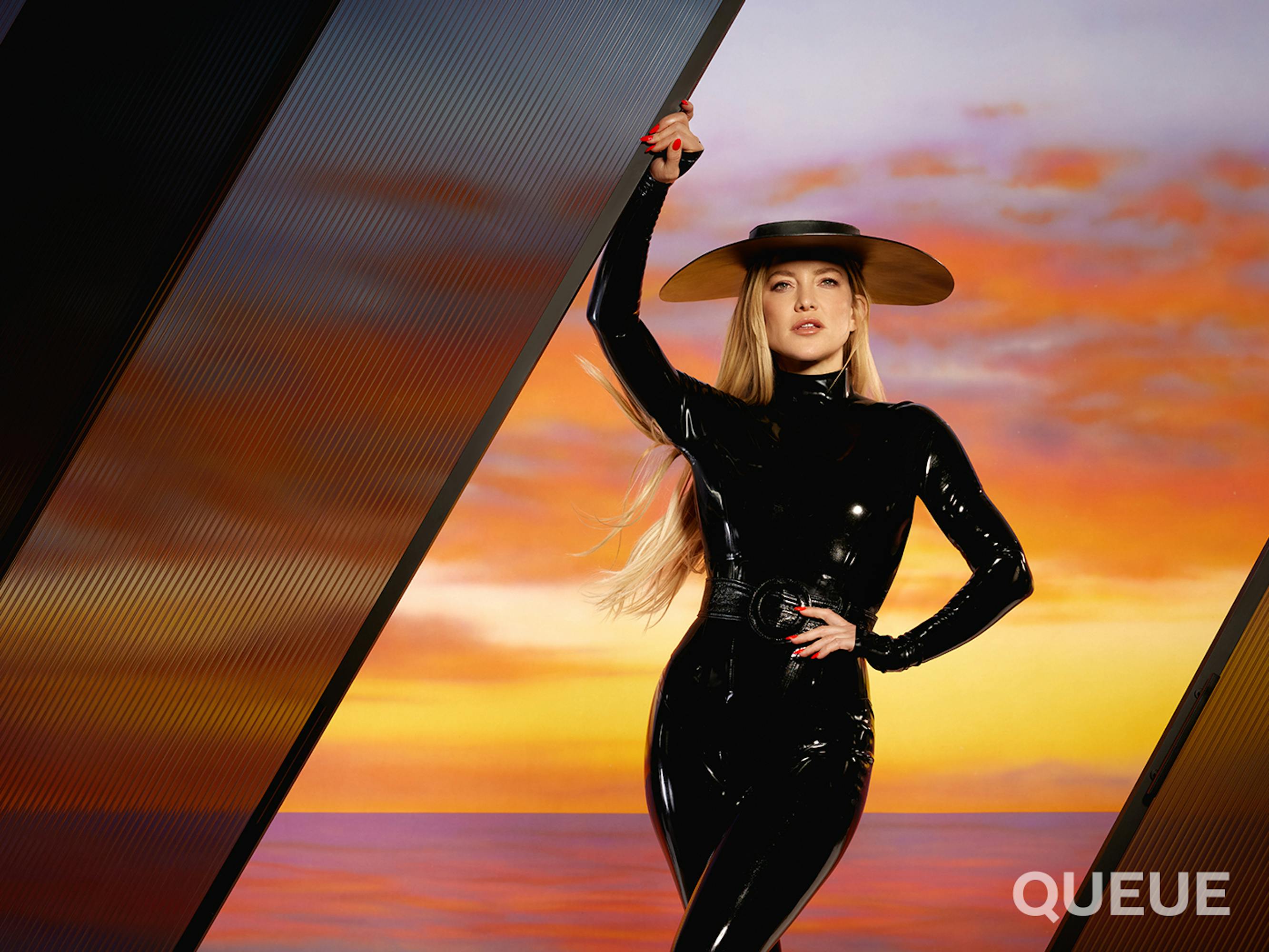 Kate Hudson wears a black latex suit and wide-brimmed hat against an orange and pink sunset.