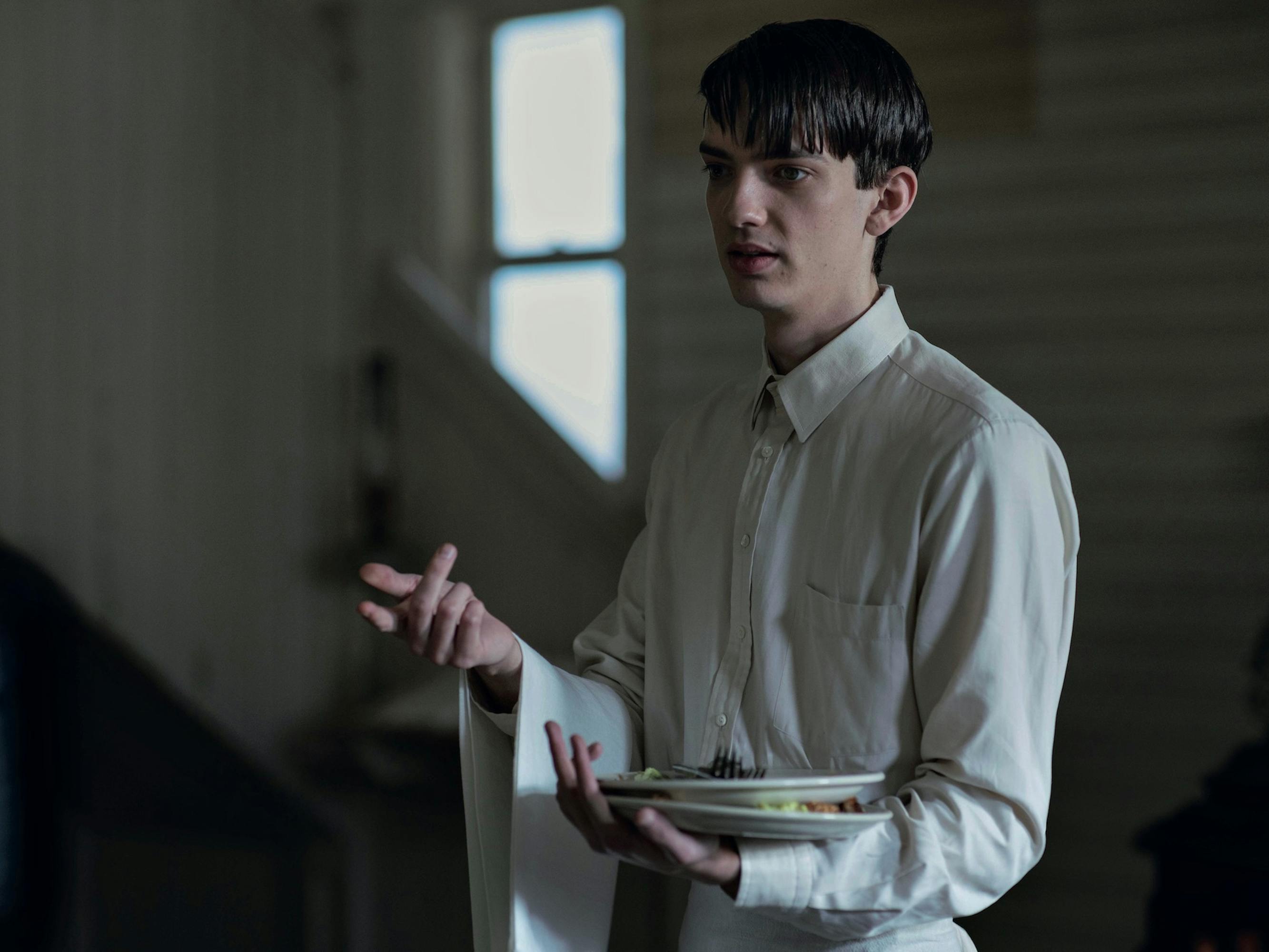 Peter Gordon (Kodi Smit-McPhee) wears a white buttoned down shirt and holds some plates in his arms.