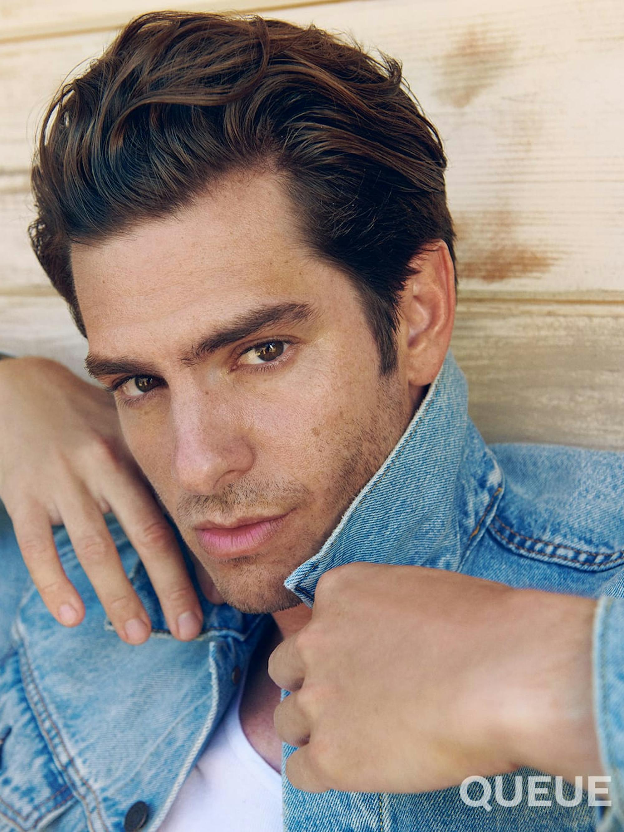 Andrew Garfield wears a light wash denim jacket pulled up to his jaw, as he stands against a wood wall.