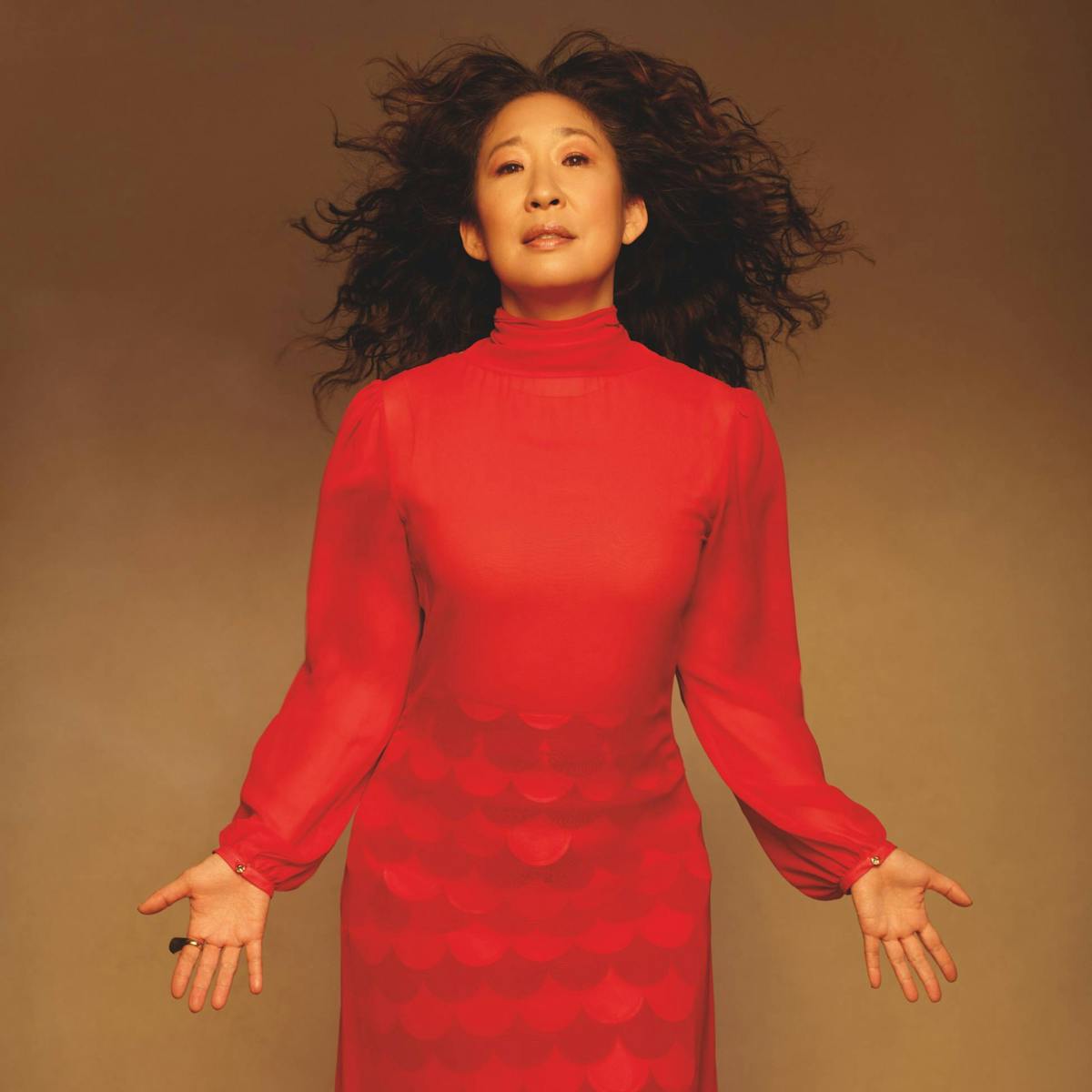Wearing a stunning long-sleeved red chiffon dress Sandra Oh moves confident & playfully in front of the camera. Her long curly hair billowing around her.