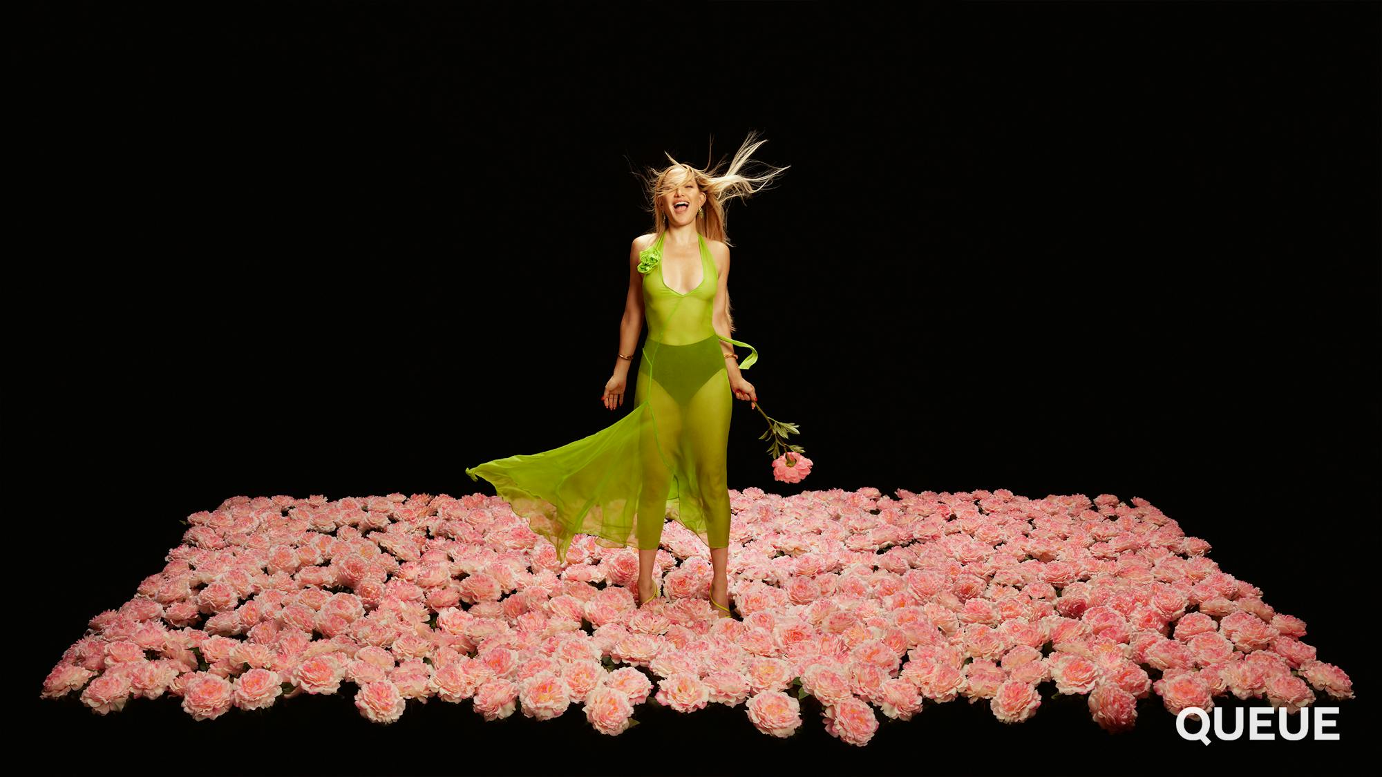 Kate Hudson wears a green sheer dress and stands in a square of flowers against a black background. 