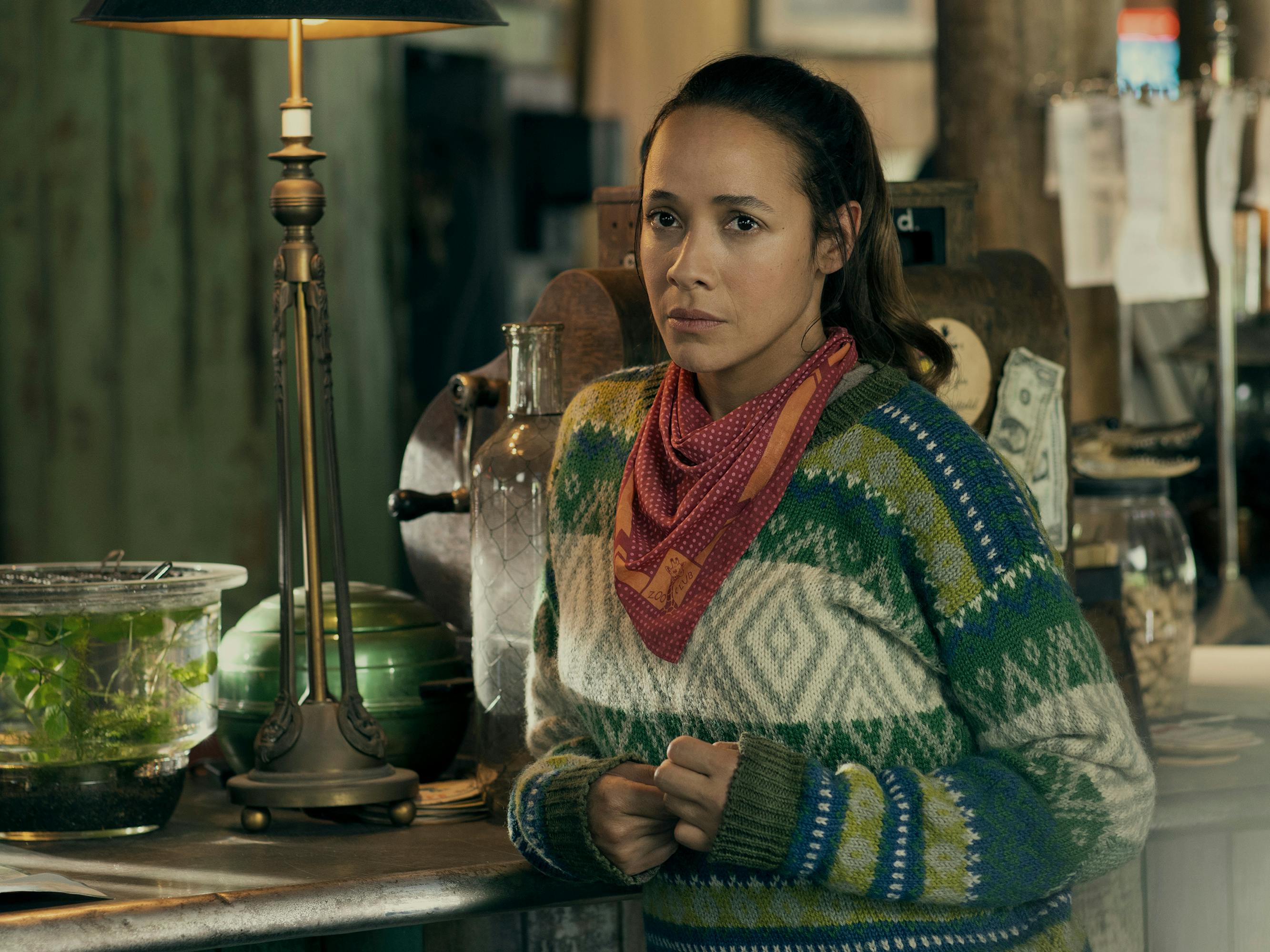 Aimee (Dania Ramirez) wears a blue-and-green sweater and a pink scarf and leans against a counter.