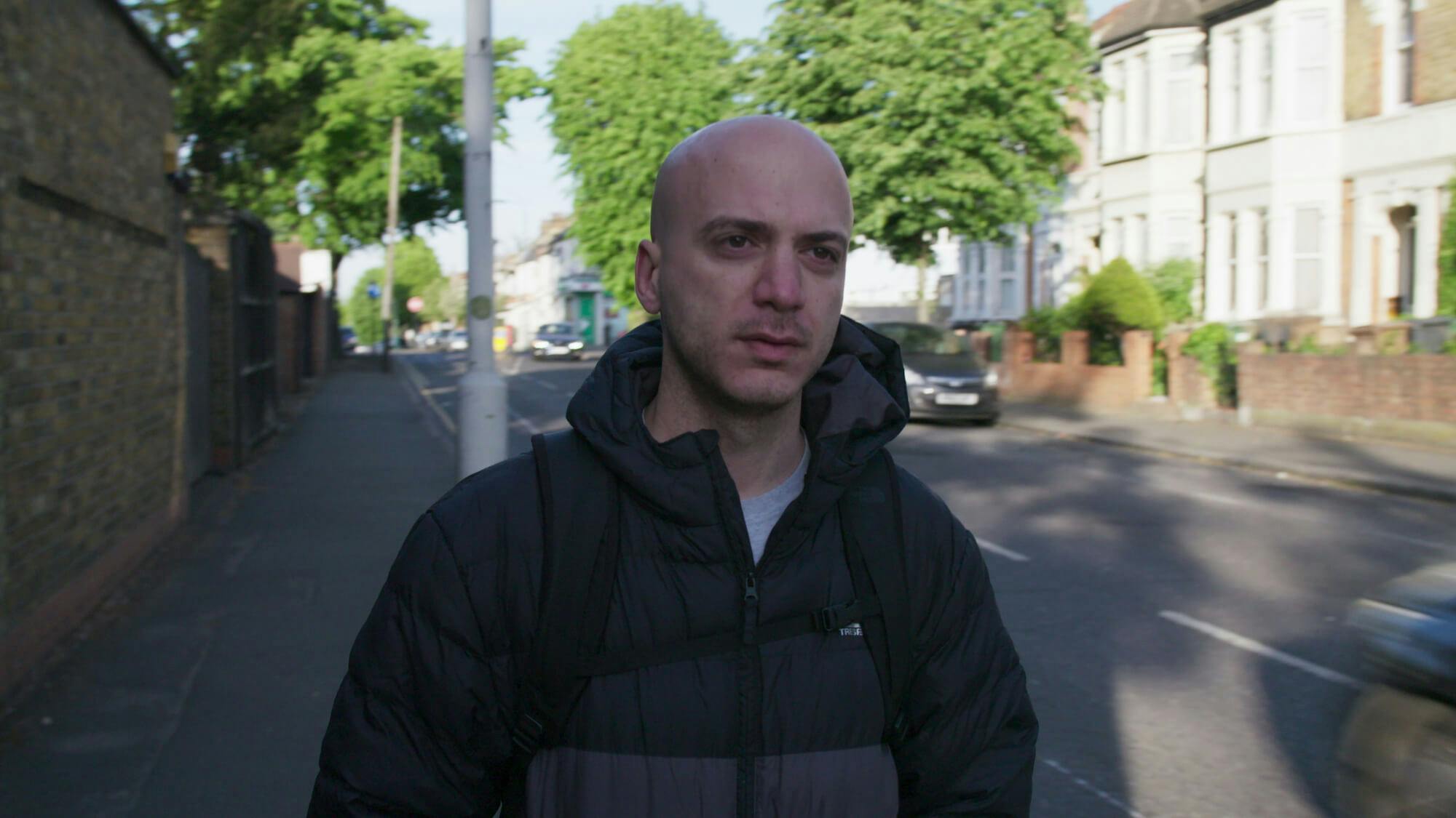 Hassan Akkad walks on a sunlit London street wearing a Northface puffer jacket.
