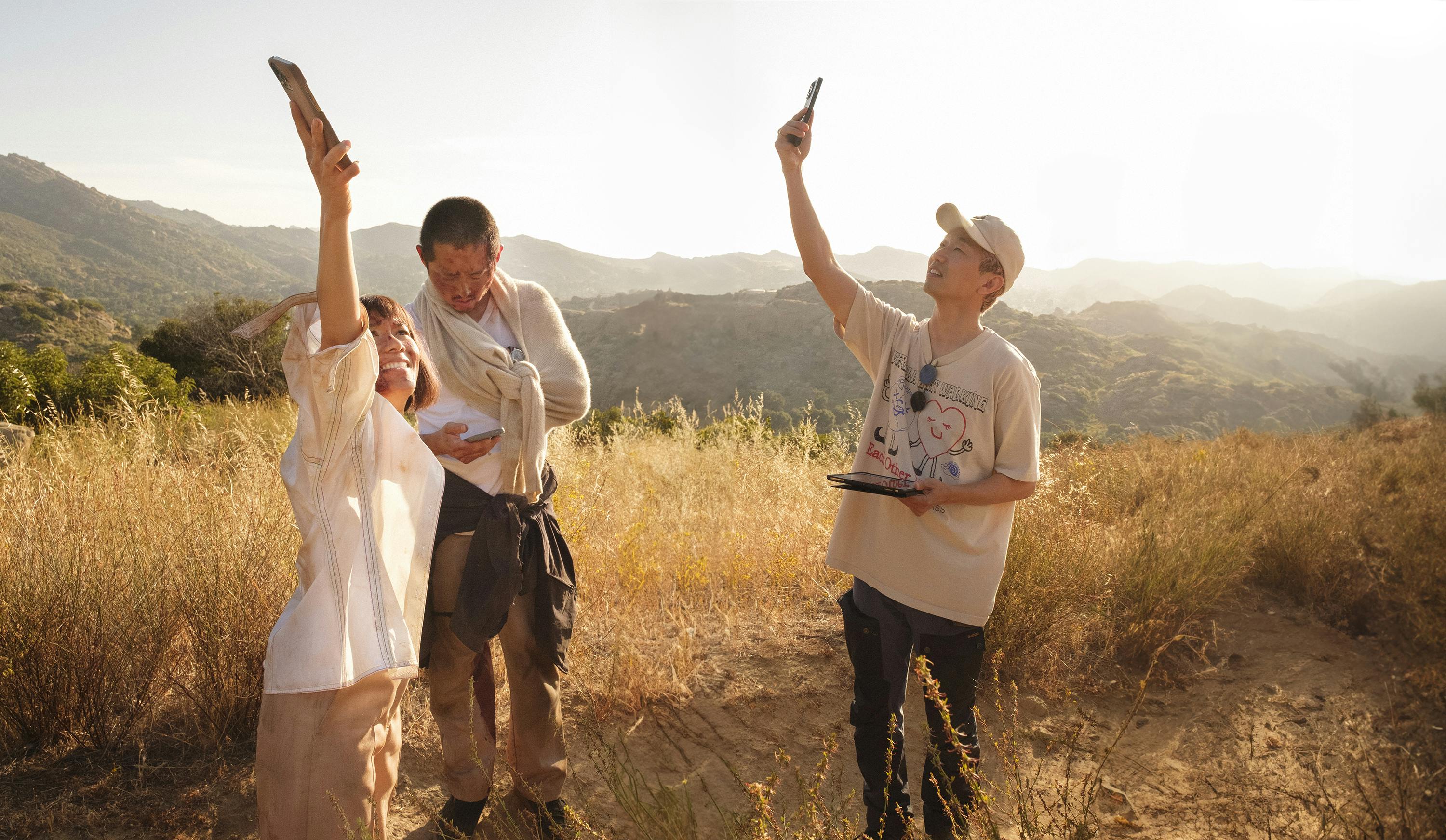 Ali Wong, Steven Yeun, and Lee Sung Jin stand in a sunny, dry field. They all wear white or beige shirts, and Lee and Wong raise phones to the sky. 