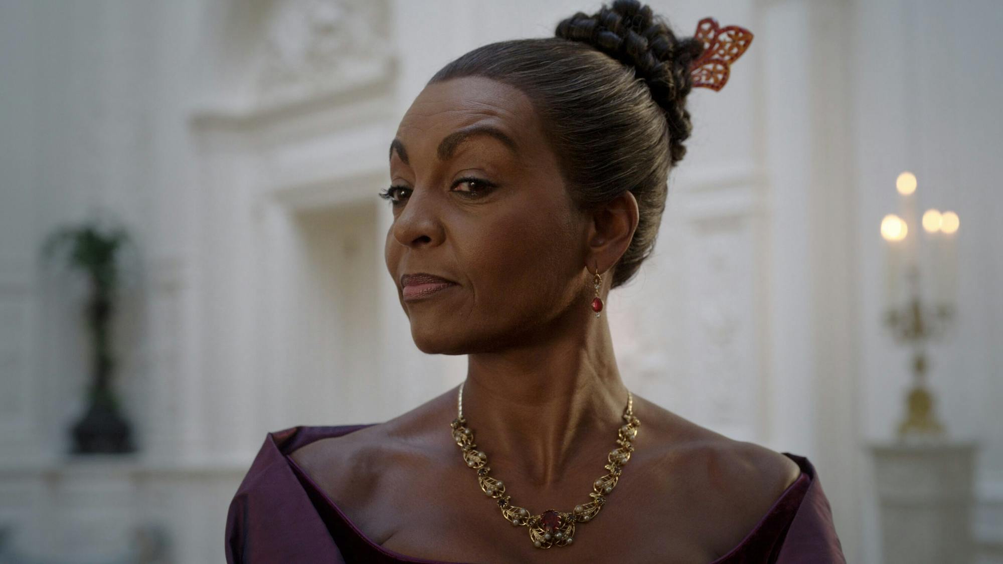 Lady Danbury (Adjoa Andoh) in an elegant shot. Her brows are raised slightly, her hair tied back with a clip. She wears a gold necklace, purple earrings, and a purple dress. The background is white, lit by a candelabra.