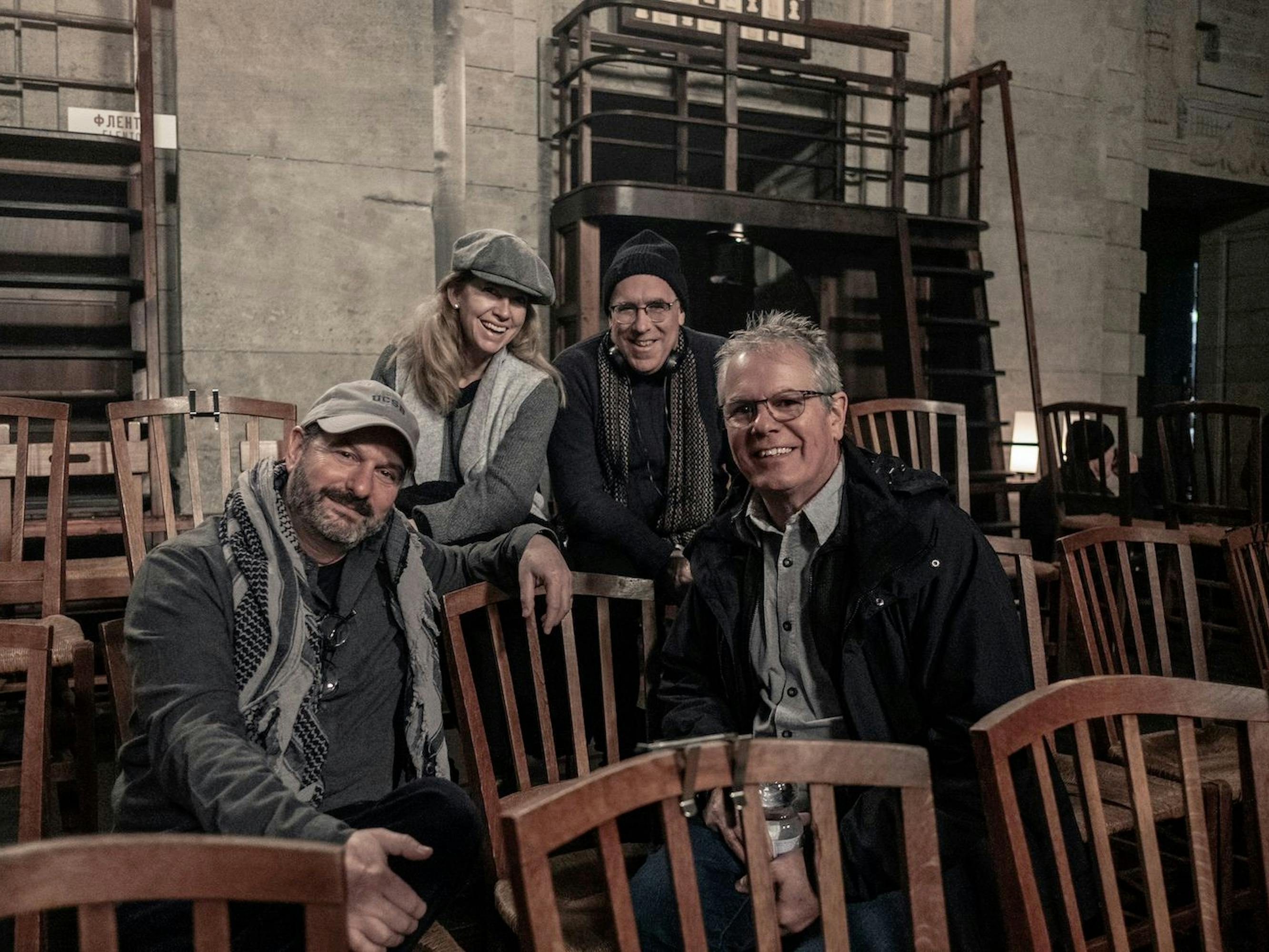 Left to right: Scott Frank, Lisa Stateman, William Horberg, Wylie Stateman. They wear an almost-uncanny mix of identical color palettes, with grey being the dominant color. The foursome sits in a crowd of wooden chairs.