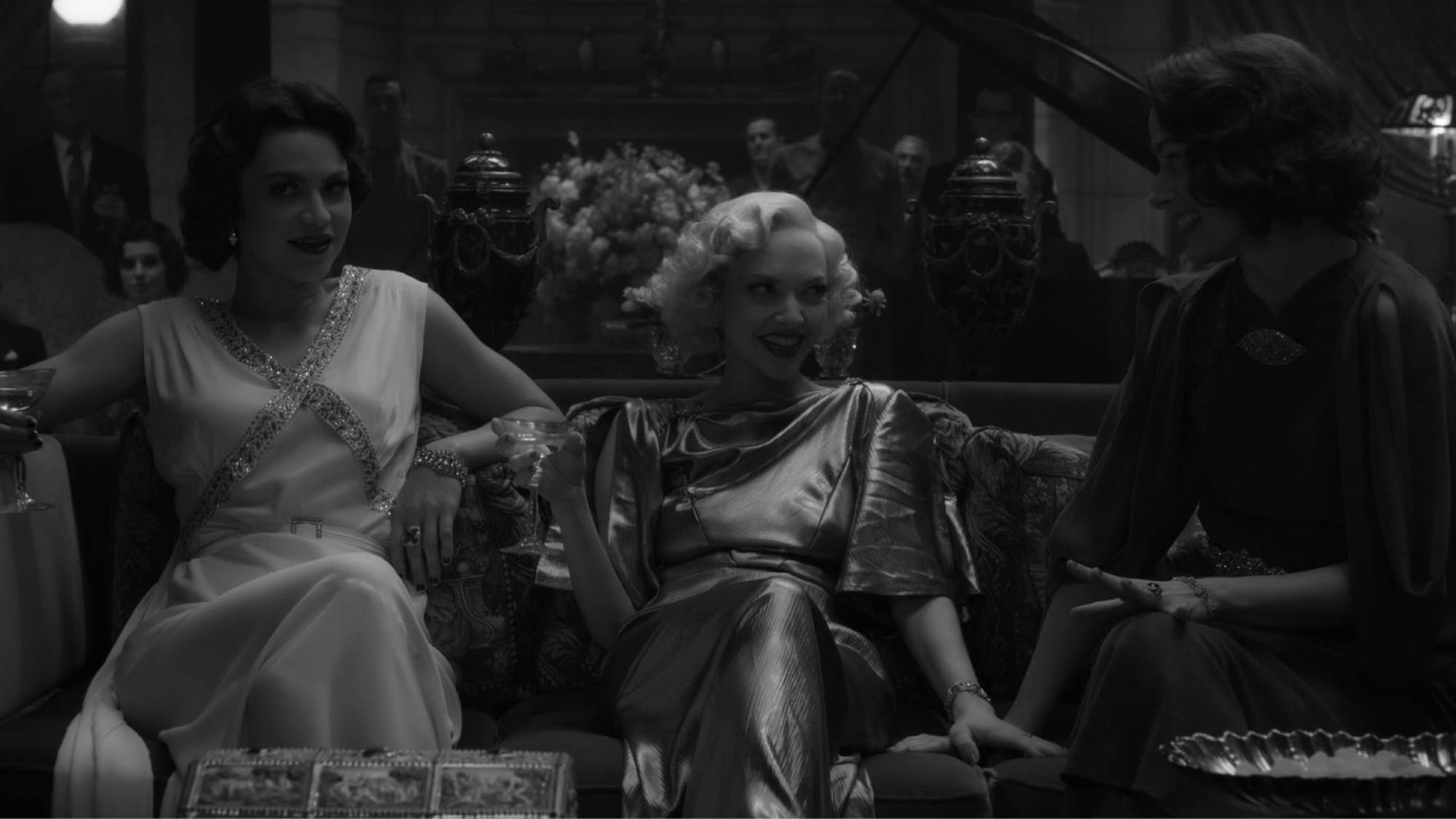 Davies is seated on a couch, talking to two women in this shot from the film. She wears a stunning gold lamé dress that falls like liquid and catches the light. She holds a cocktail. Other party guests are visible in the background, between ornate urns and flower arrangements.