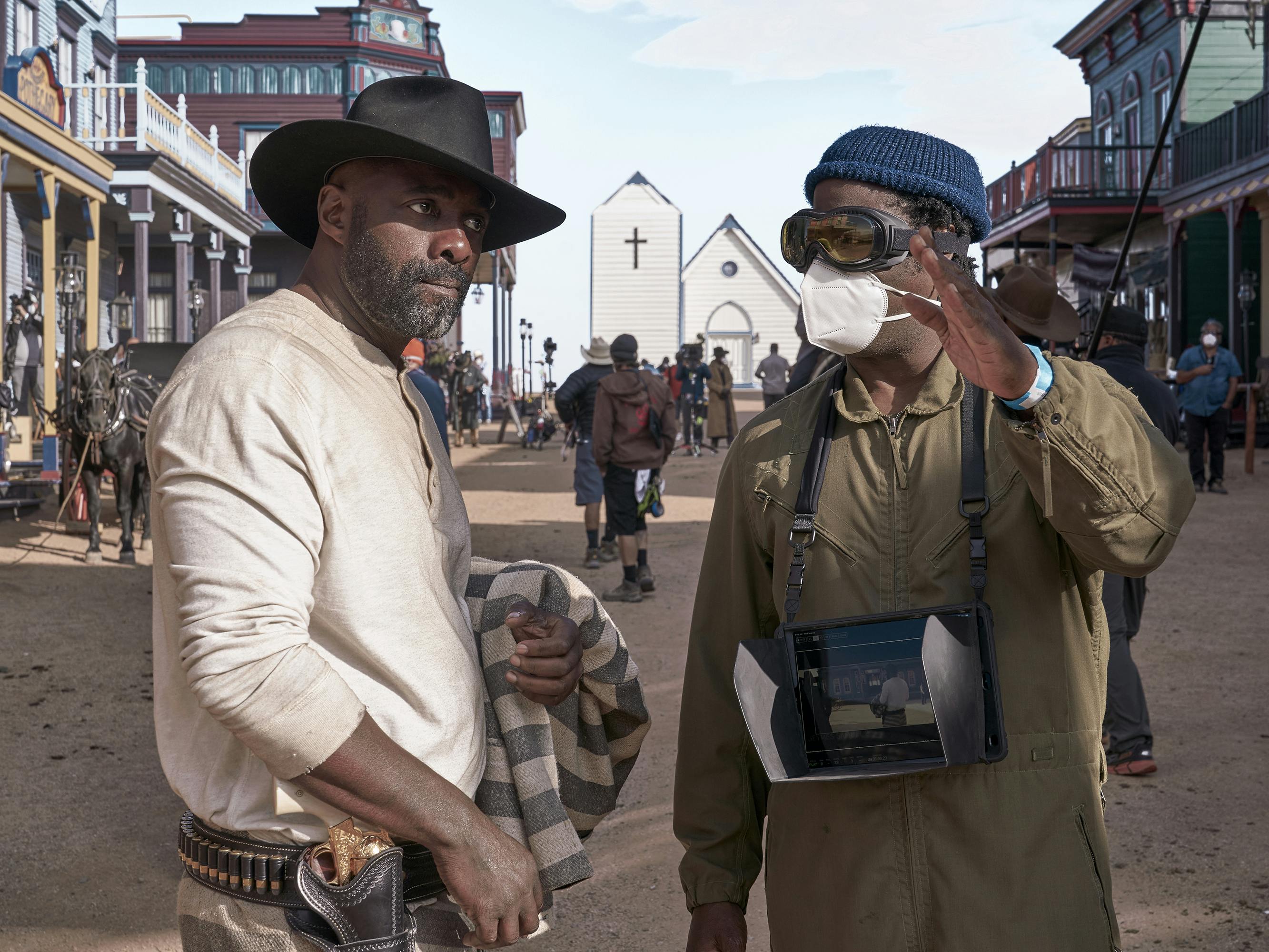 Idris Elba and Jeymes Samuel stand in the middle of the street. Elba wears a white shirt and dark wide-brimmed hat. Samuel wears a green khaki jumpsuit, goggles, and a blue hat. 