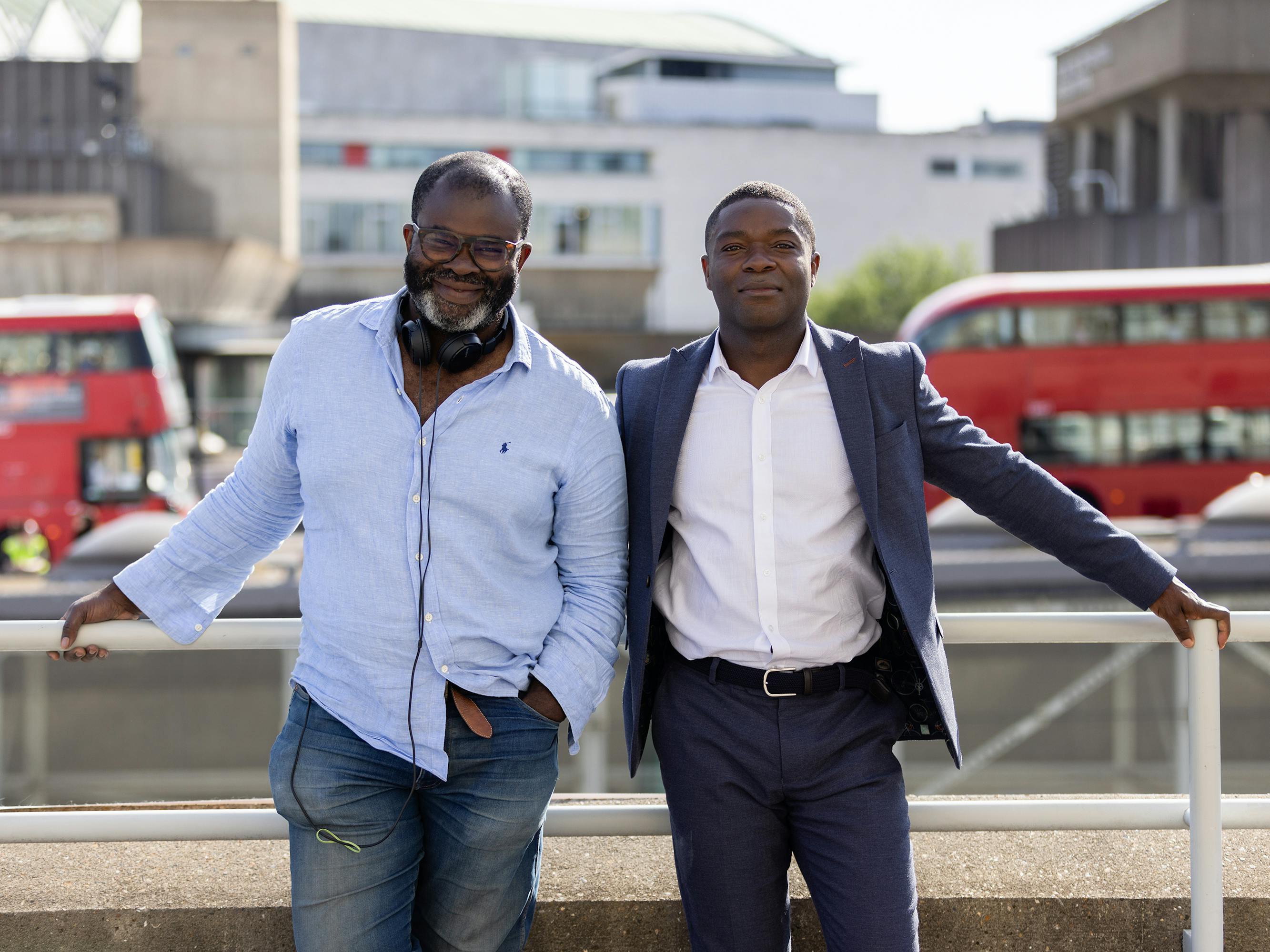 Misan Harriman and David Oyelowo stand together on a bridge. Misan wears a chambray shirt and jeans. David wears a navy suit with a white dress shirt.
