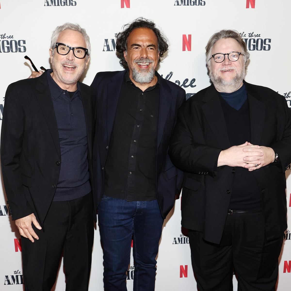 Alfonso Cuarón, Alejandro Iñárritu, and Guillermo del Toro stand together on a red carpet. The step-and-repeat background features the Netflix icon, and 'the three Amigos' in faded black.