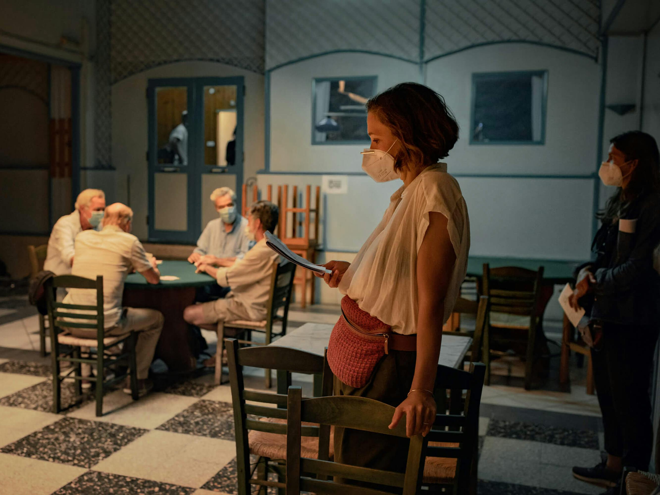 A behind the scenes shot of The Lost Daughter cast and crew. Four men sit around a green-tableclothed table. There’s a woman in the foreground wearing a pink fanny pack and white blouse. All of the figures in this shot wear masks.