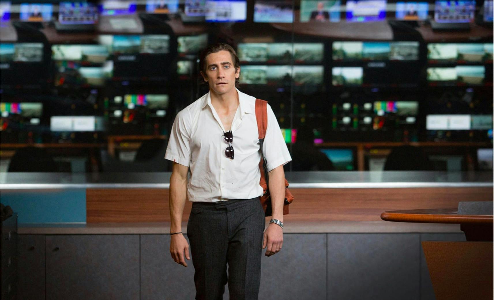 Louis Bloom (Jake Gyllenhaal) in Nightcrawler (2014) wears a white shirt, dark pants, and an orange backpack. There are many screens in the background.