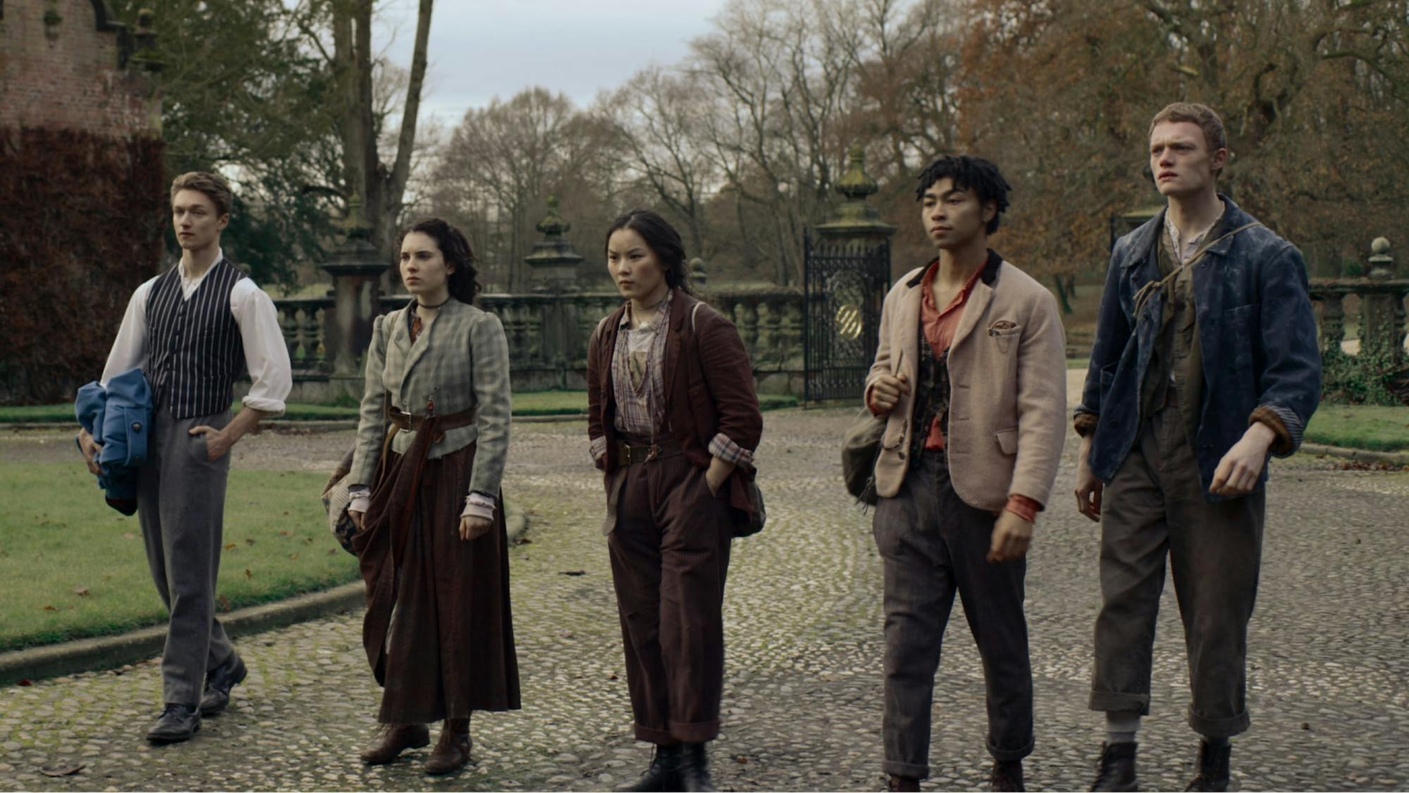 The cast walks in a line through a cobblestone courtyard in this shot from the series. Behind them, the courtyard opens into grounds, with a line of trees visible, their leaves in reds and oranges. The characters are decked out in steampunk chic: baggy trousers, plaids, stripes, vests, and blazers — all the worse for wear.