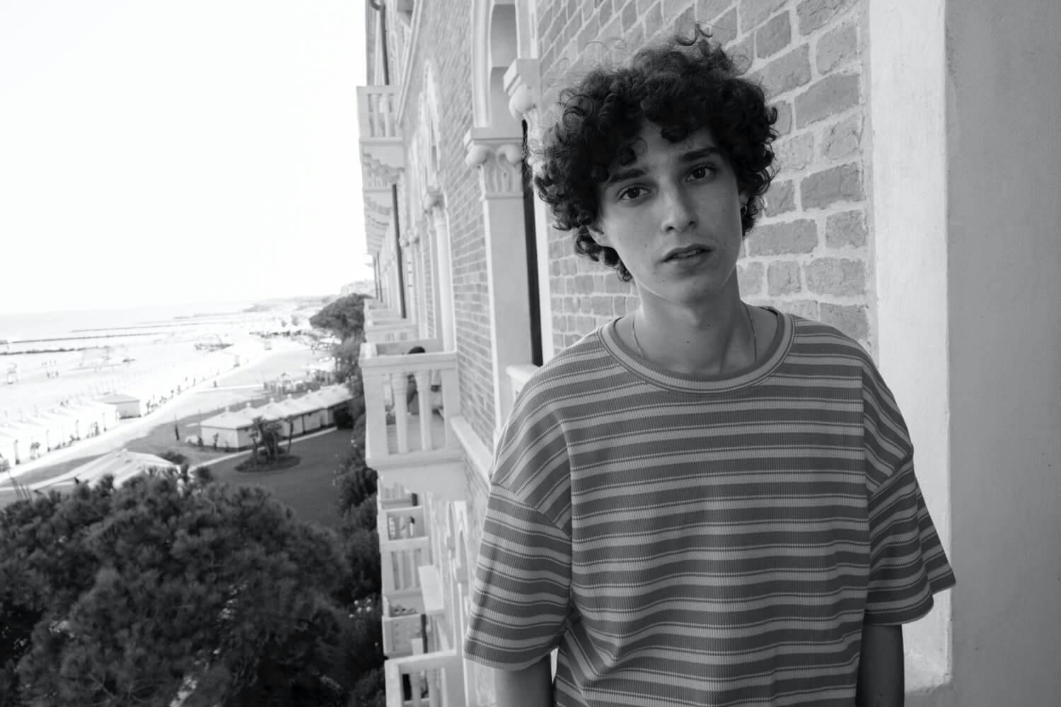 Filippo Scotti at the Venice Film Festival, 2021 wears a striped tshirt and stands on a balcony with a beach in the background.