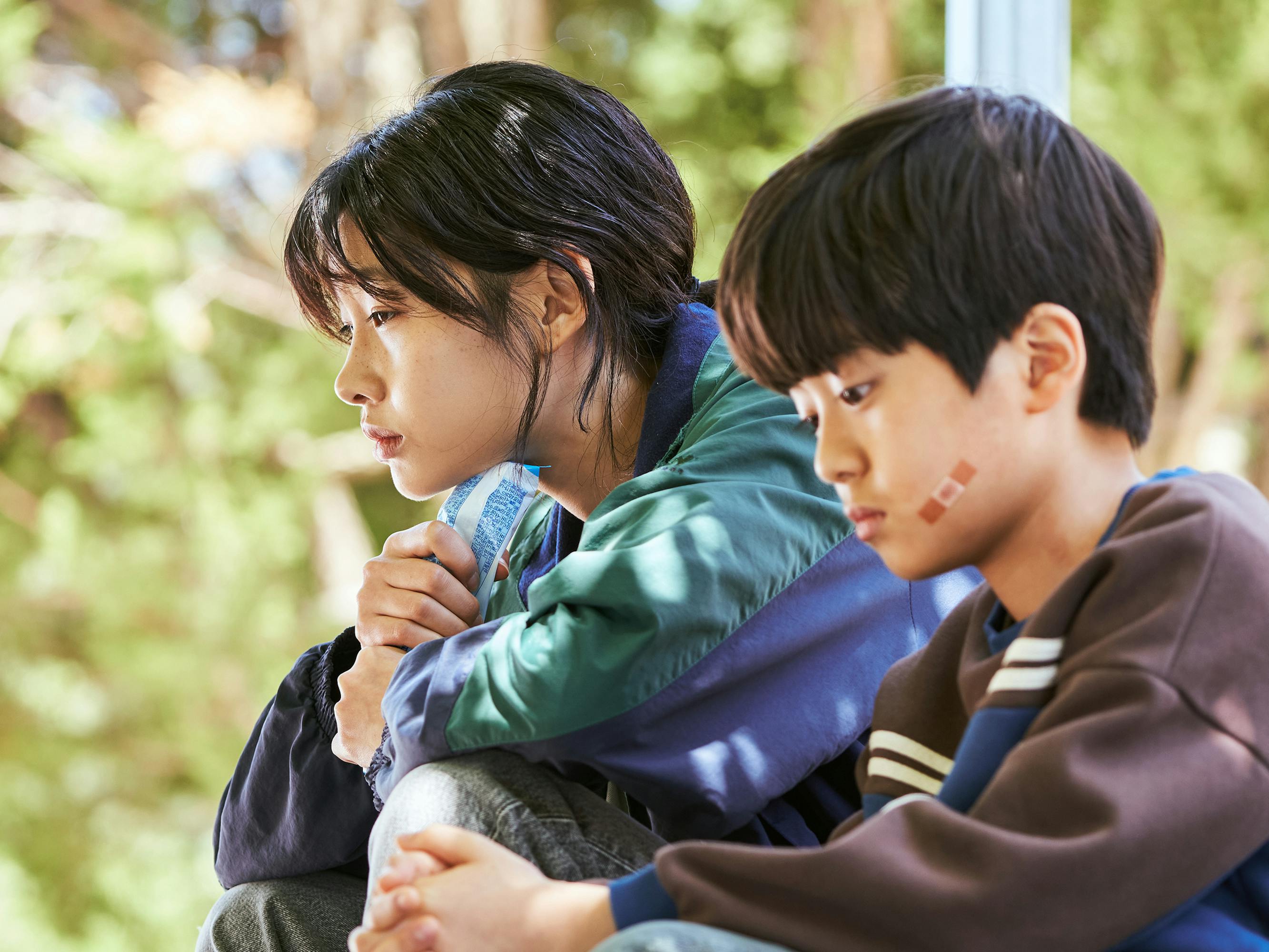 Kang Sae-byeok (Jung Ho-yeon) and Kang Cheol (Park Si-Wan) sit outside together. Sae-byeok wears a green and blue top. Kang-cheol wears a brown striped sweatshirt.