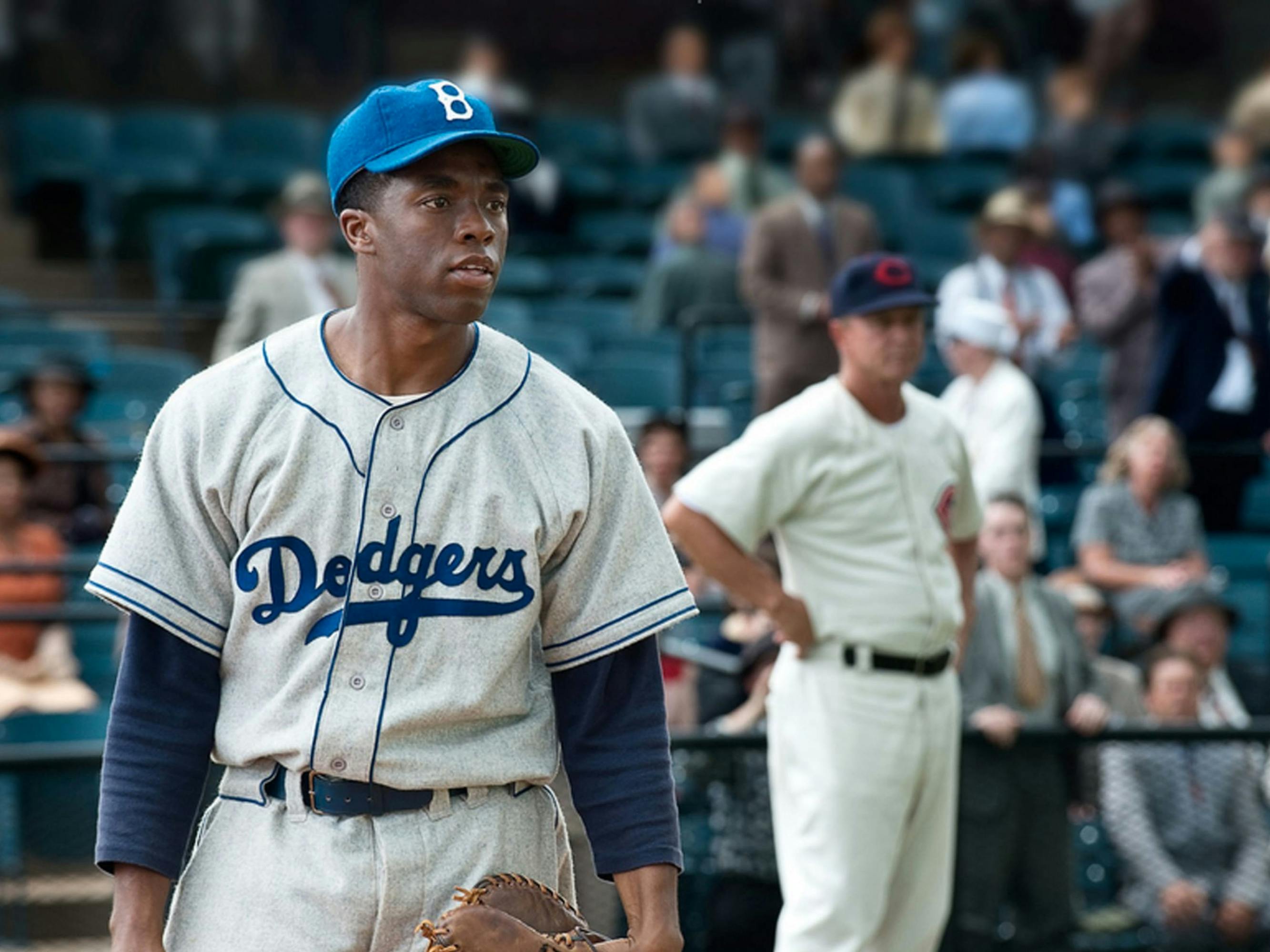 Boseman as Jackie Robinson. He wears a white Dodgers uniform with blue stitching and a blue Dodgers hat with a B for Brooklyn. He also carries a mit. In the background there are spectators in the stands and players from the opposite team.