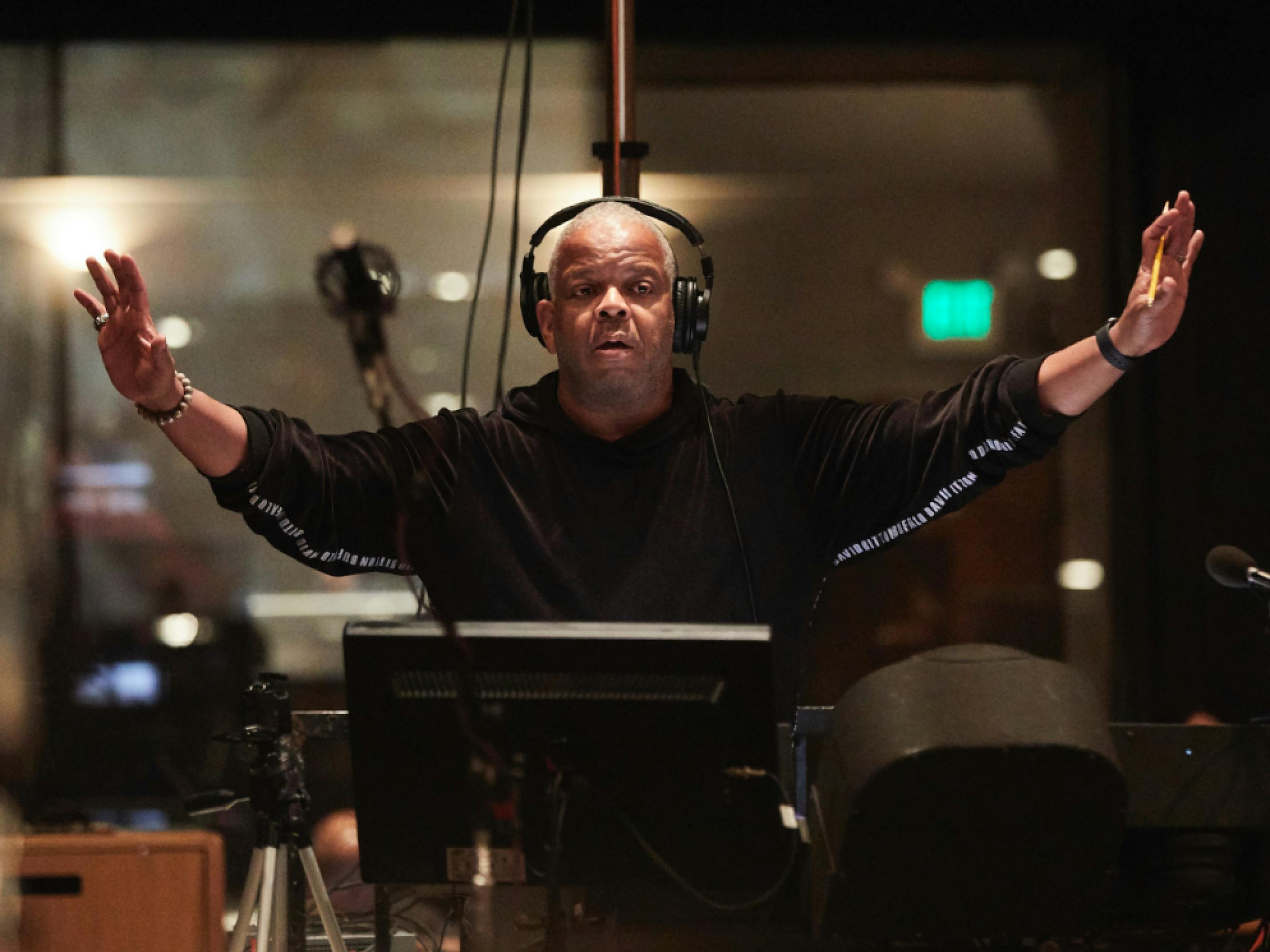 Composer Terence Blanchard is at his music stand with his arms raised up as he conducts. He has on large, over-the-ear headphones and a black hoodie.