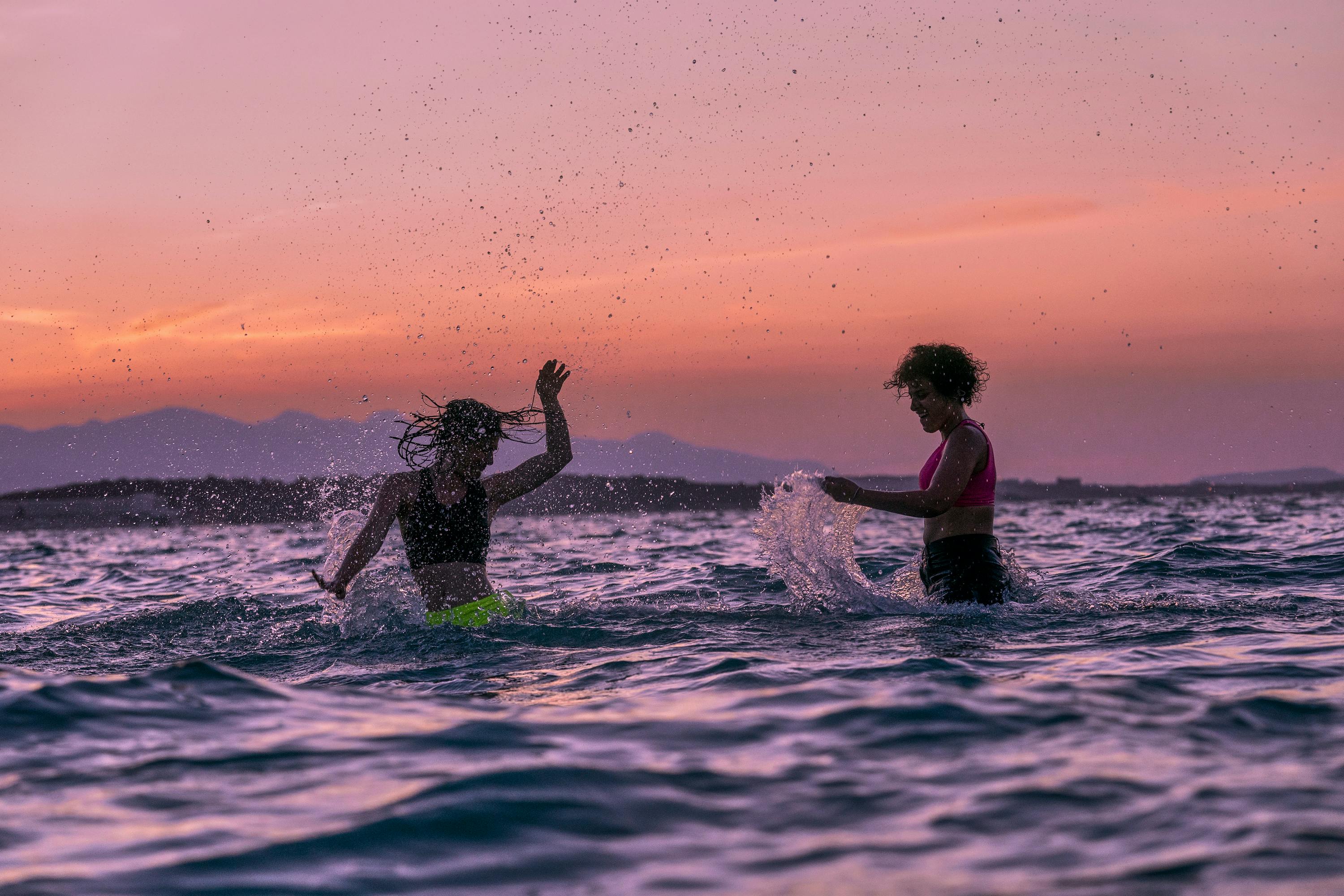 Manal and Nathalie Issa play in the water, lit by a pink and orange sunset.