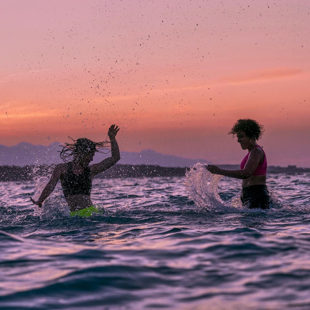 Manal and Nathalie Issa play in the water, lit by a pink and orange sunset.