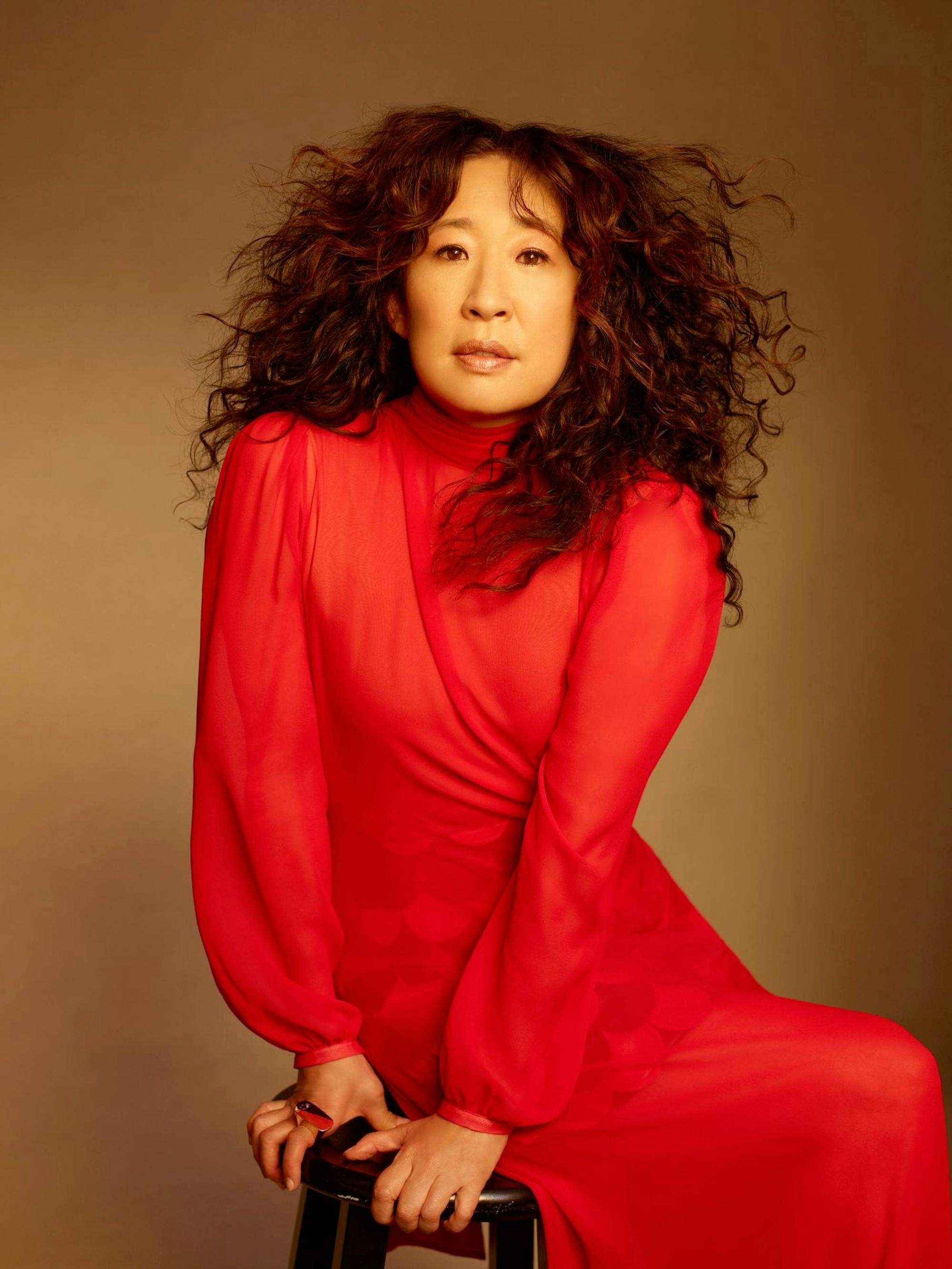 Sandra Oh wears a red dress and sits on a stool looking directly at the camera. Her hair is wild and her face is lit by a warm light.