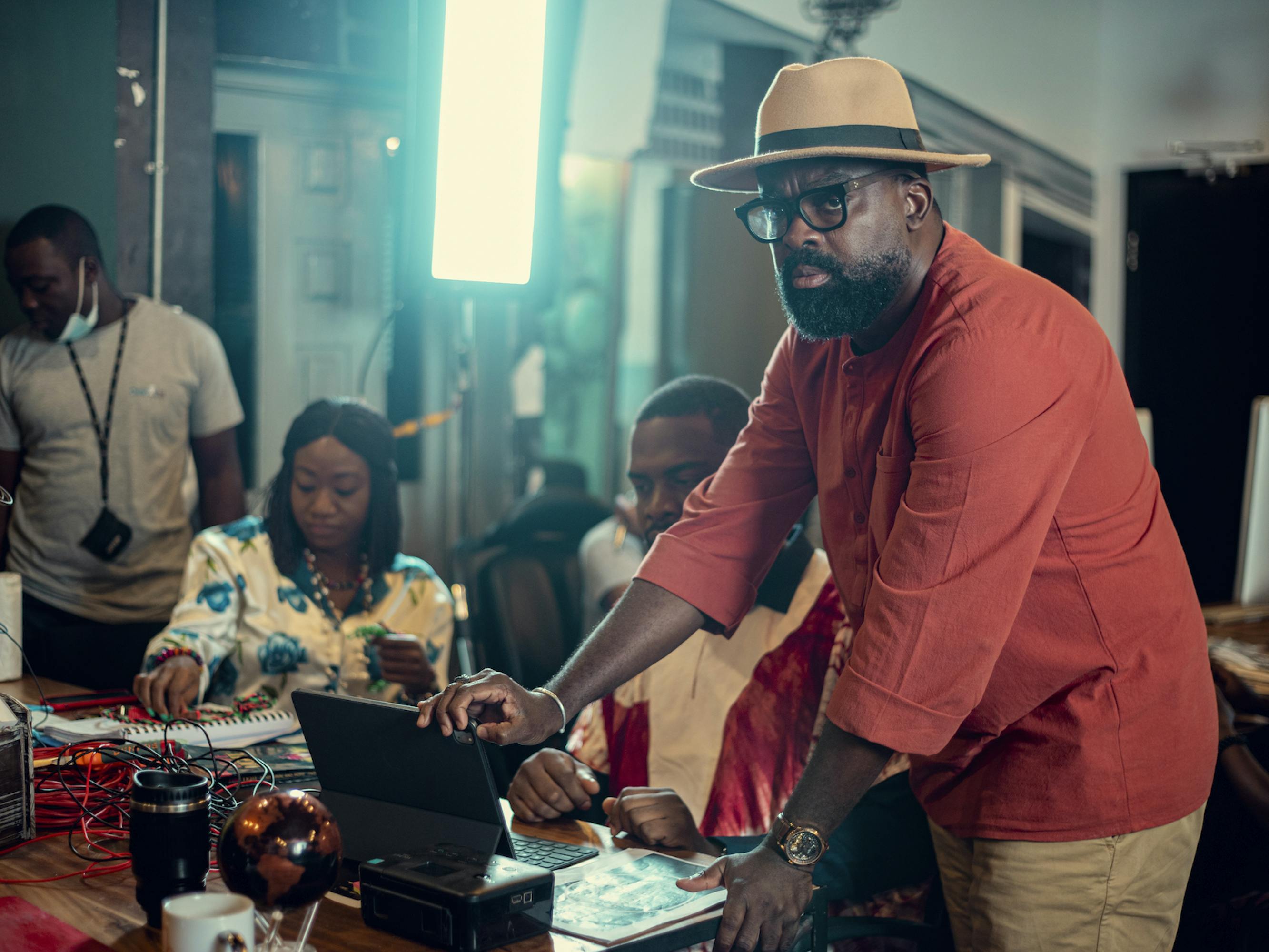 Kunle Afolayan stands around a table with other crew members. He wears a red shirt, khakis, and a straw hat with a black ribbon. 