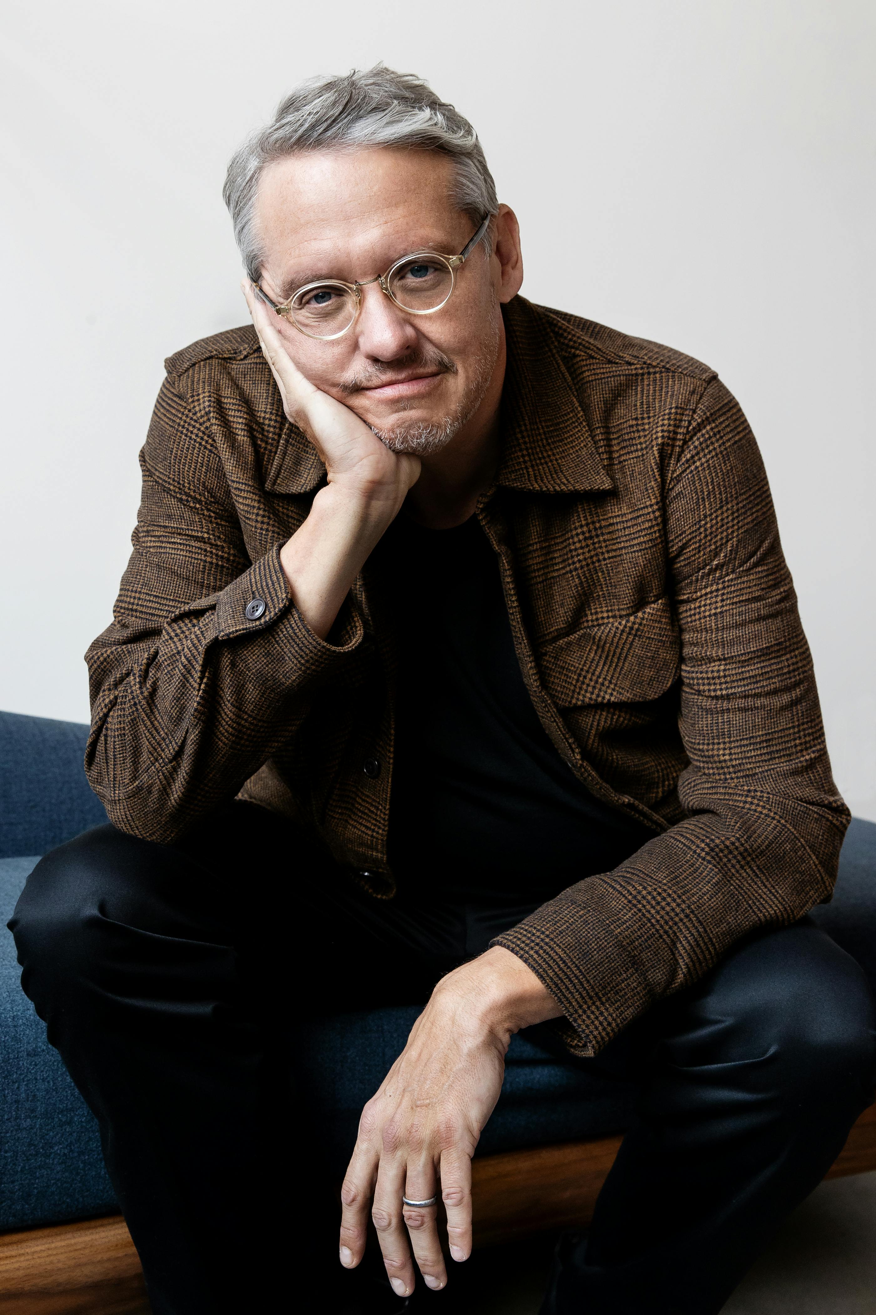 Adam McKay wears jeans and a brown jacket with his chin resting on his palm.