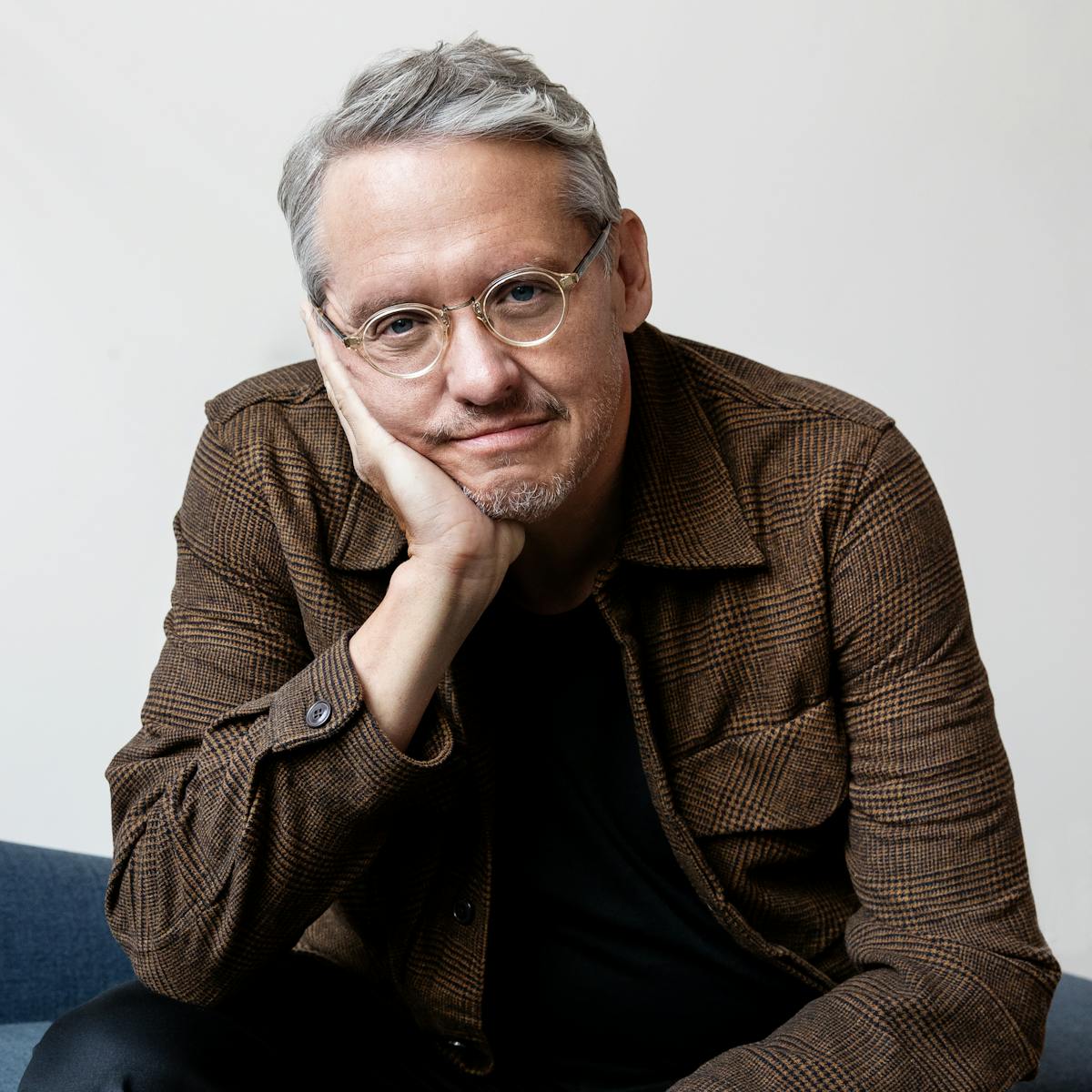 Adam McKay wears jeans and a brown jacket with his chin resting on his palm.