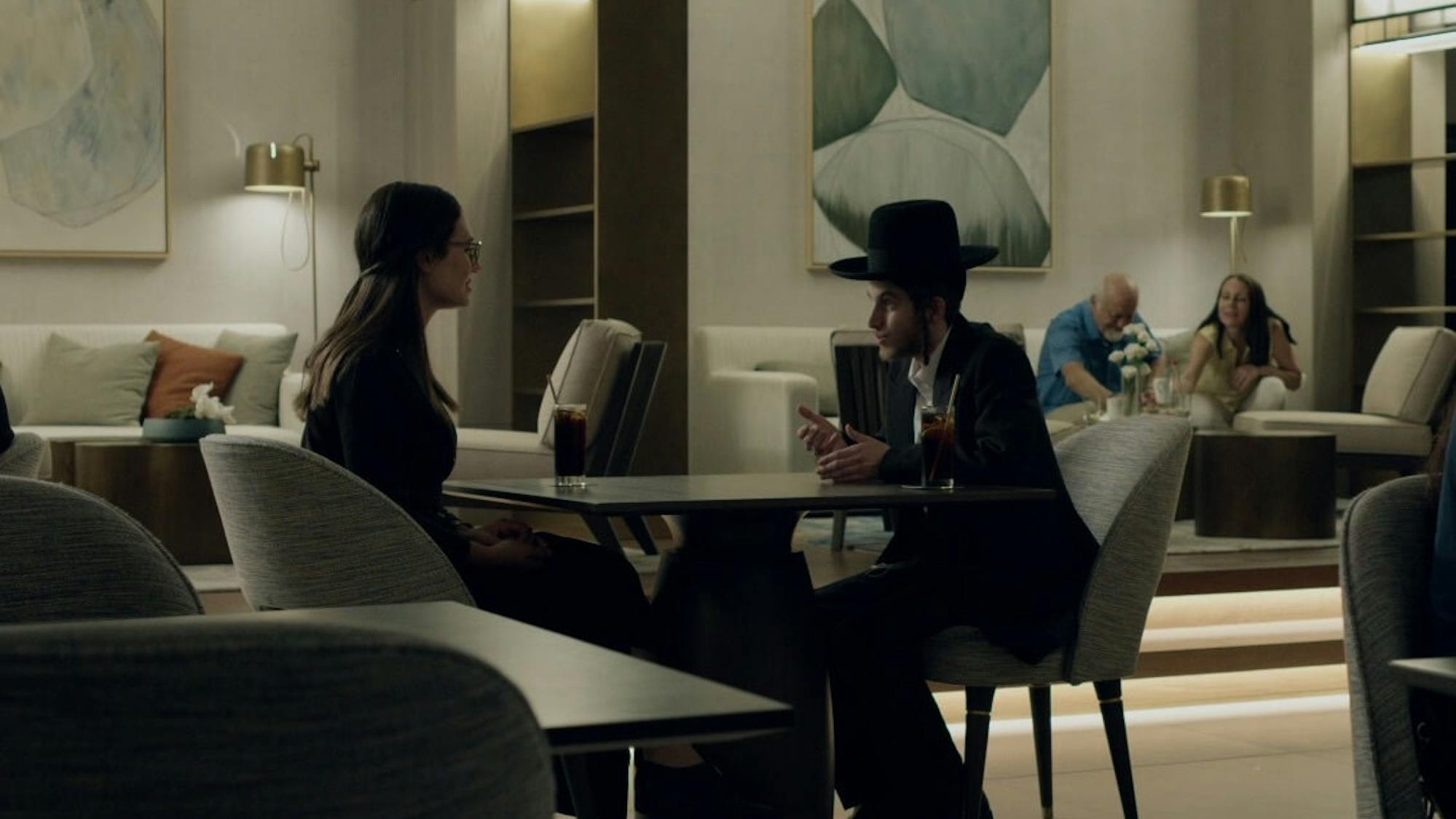 This shot is full of shadowy figures sitting at tables or couches. In the middle are a man and woman at a table talking over two sodas. The scene looks more modern than some of the other shots from Shtisel.