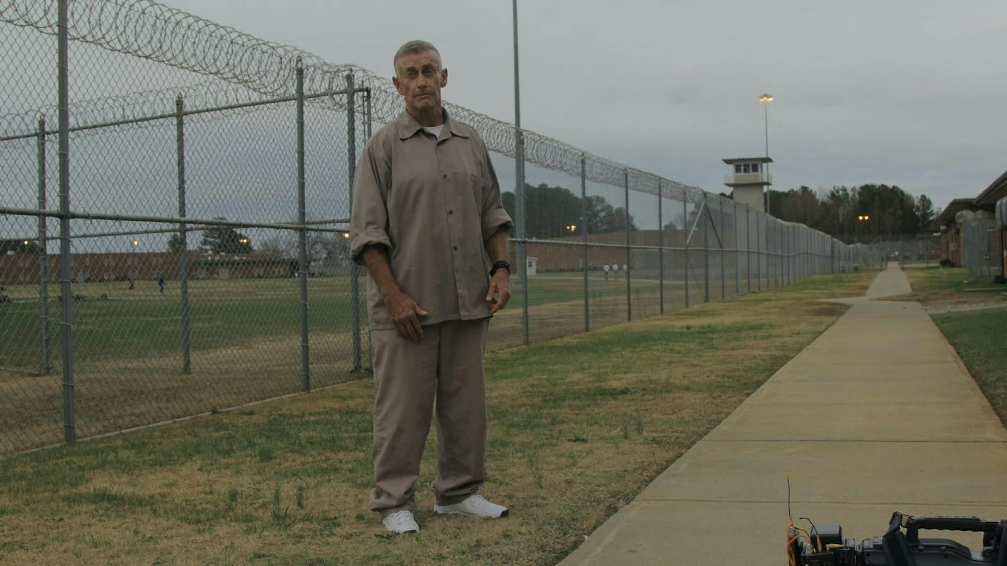 A shot from The Staircase. Michael Peterson wears tan prisonwear and stands on the grass between a sidewalk and a barbed fence.