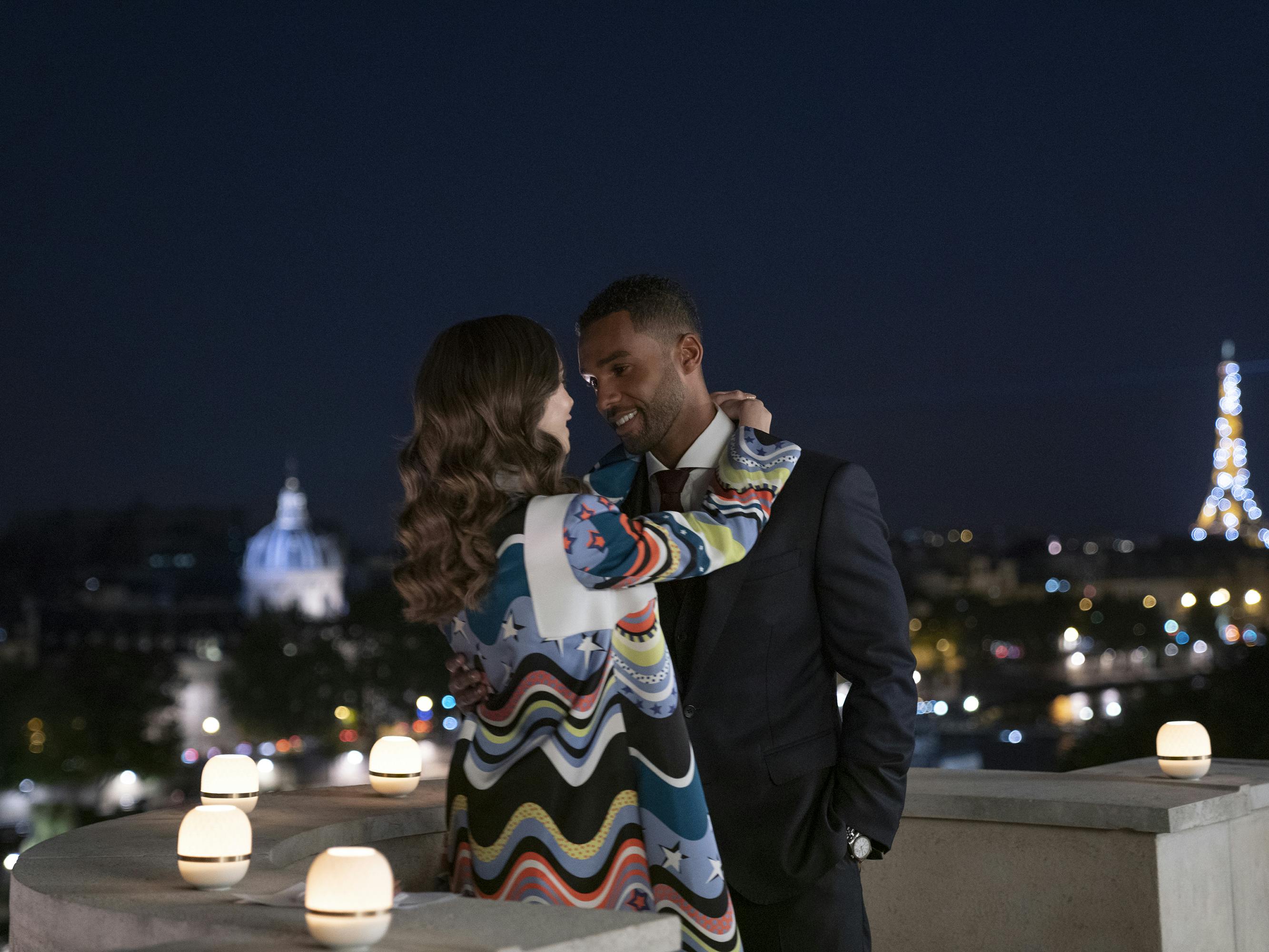 Emily Cooper (Lily Collins) embraces Alfie (Lucien Laviscount) while the Eiffel Tower shimmers in the background. Emily wears a colorful jacket and alfie