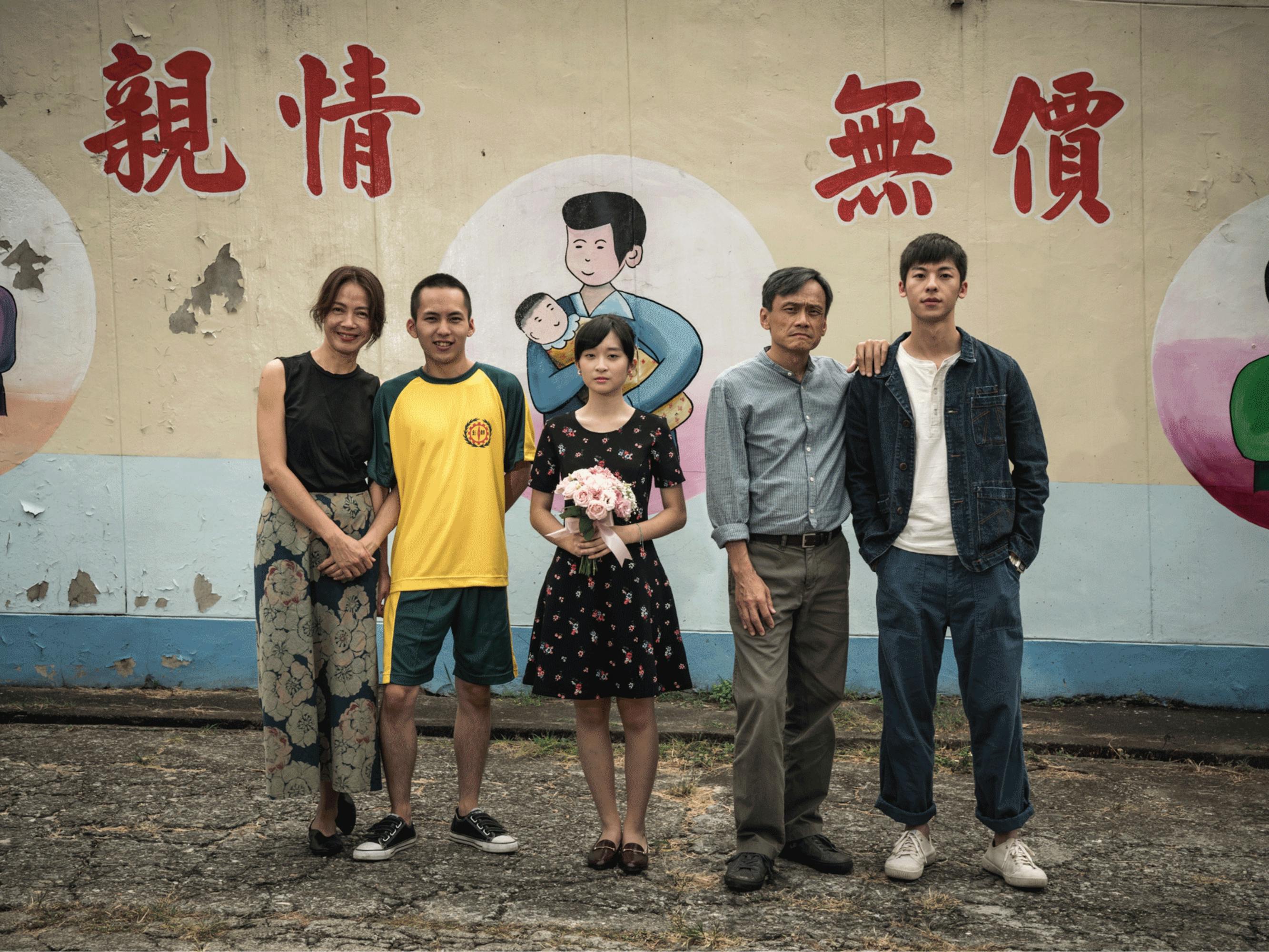 In this still from the film, the actors stand in front of a painted mural showing simple drawings of women holding infants. Wu Tai-ling, in the center, holds a bouquet of pink flowers. Samantha Ko and Wu Chien-ho stand in a pair on the left. Chen Yi-wen places a hand on Hsu Kuang-han’s shoulder. 